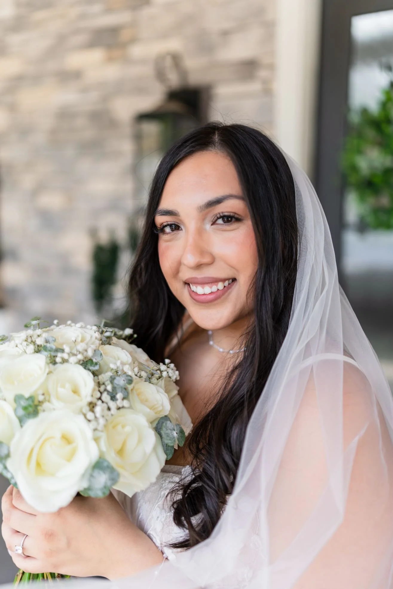 A smiling bride with long dark hair, wearing a white dress and veil, holding a bouquet of white roses and greenery.