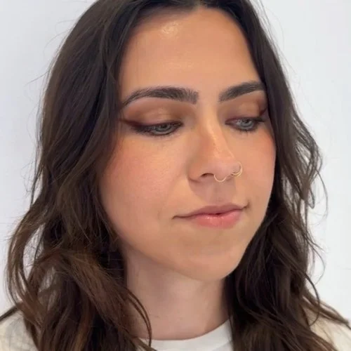 Close-up of a woman with dark wavy hair, light makeup, and a nose ring, looking downward.