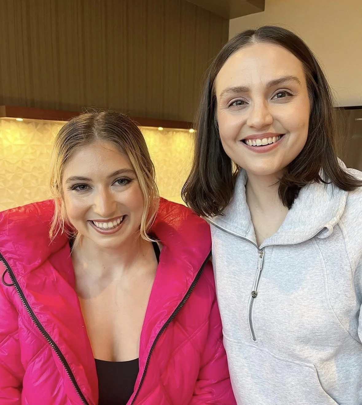 Two women smiling, one wearing a pink jacket and the other wearing a light gray hoodie, in an indoor setting with warm lighting.
