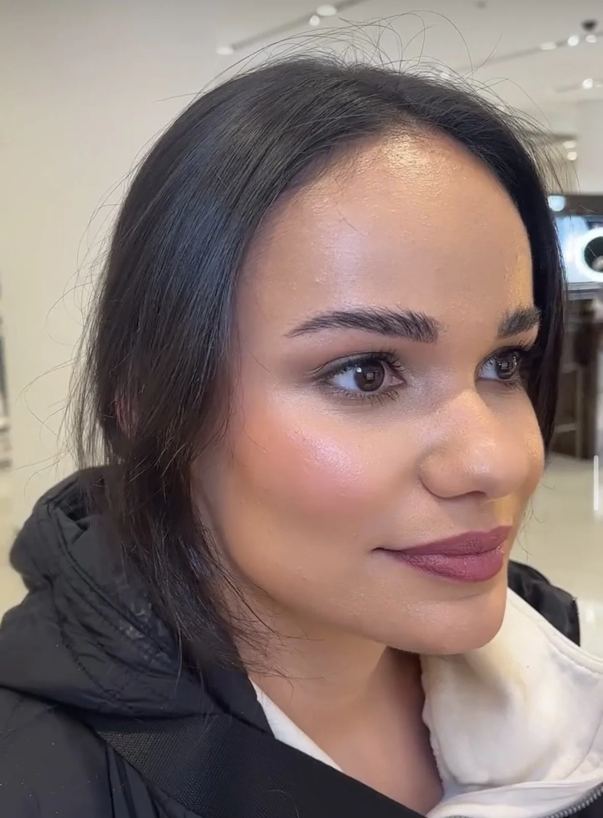 Close-up of a woman with shoulder-length dark hair, wearing makeup with neutral tones, and dressed in a black jacket and white hoodie.