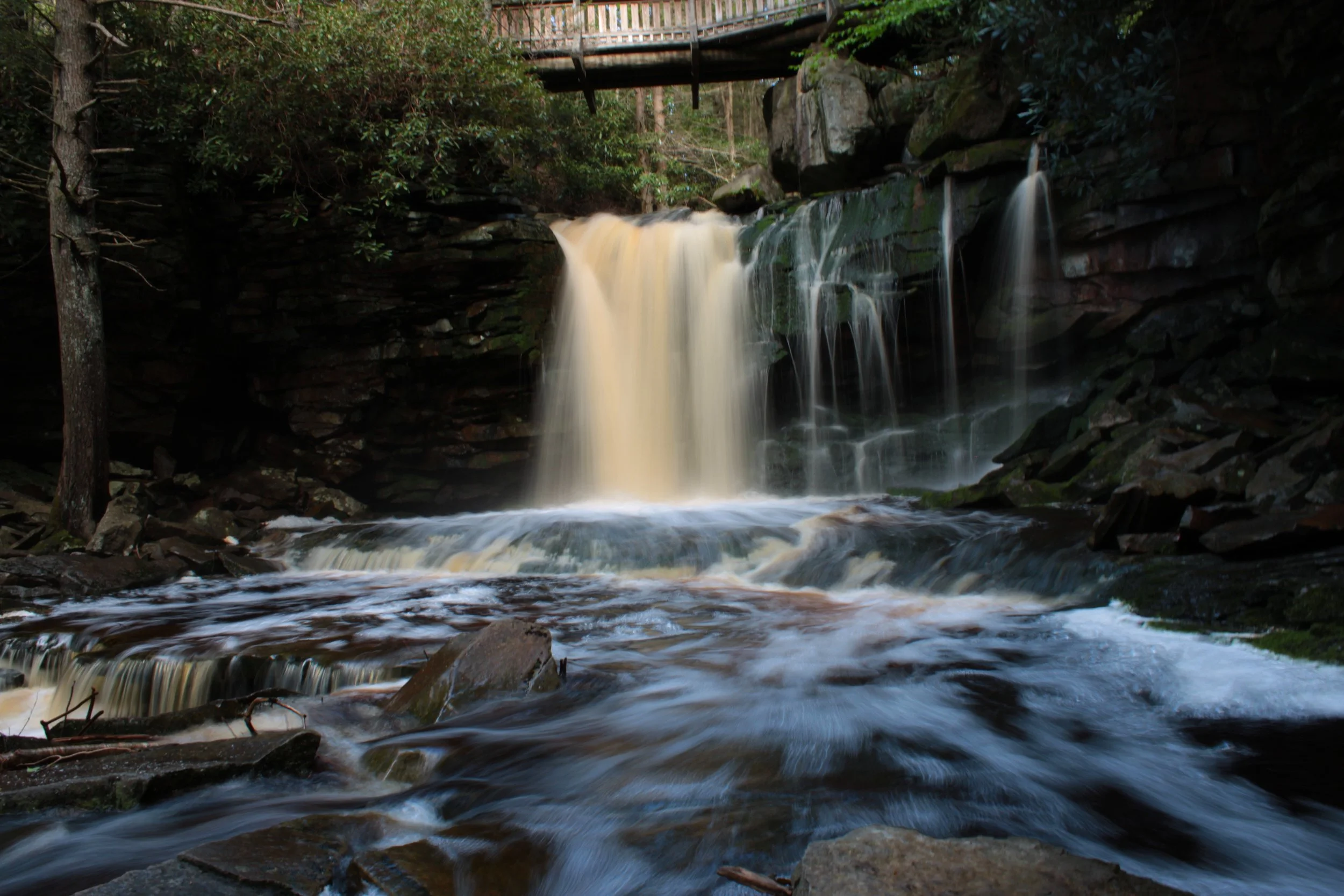 A reddish brown waterfall flowing over rocks surrounded by forest with a wooden bridge above.