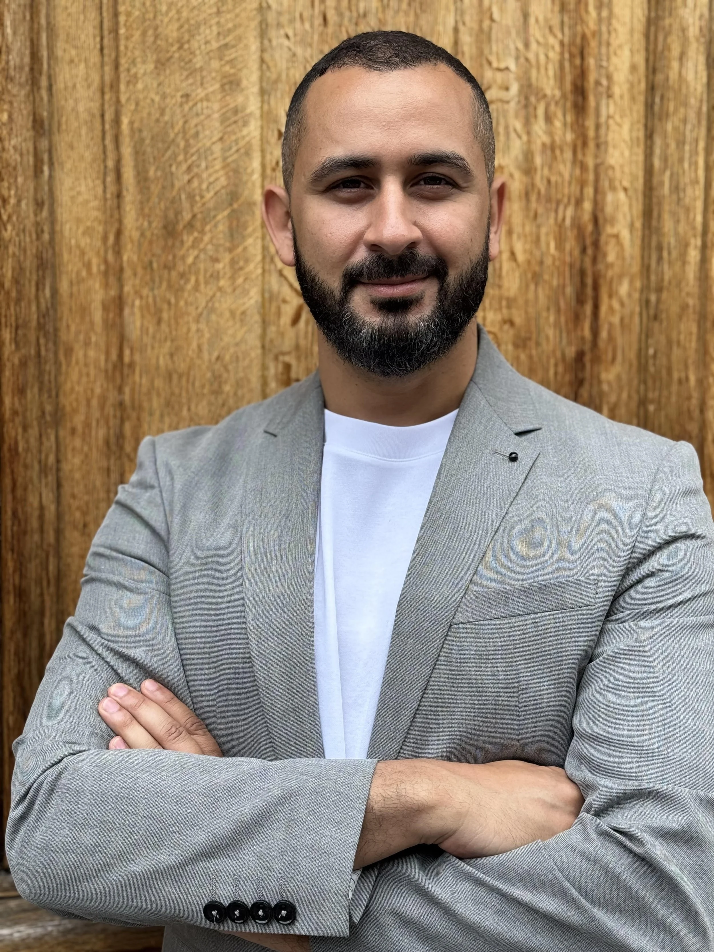 Homme souriant avec une barbe, vêtement gris, bras croisés, fond en bois.