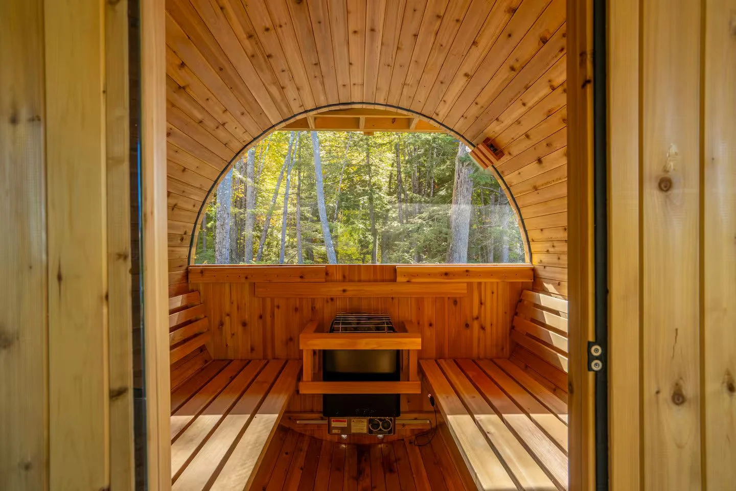 Interior view of a small wooden sauna with a semi-circular window showing a forest outside.