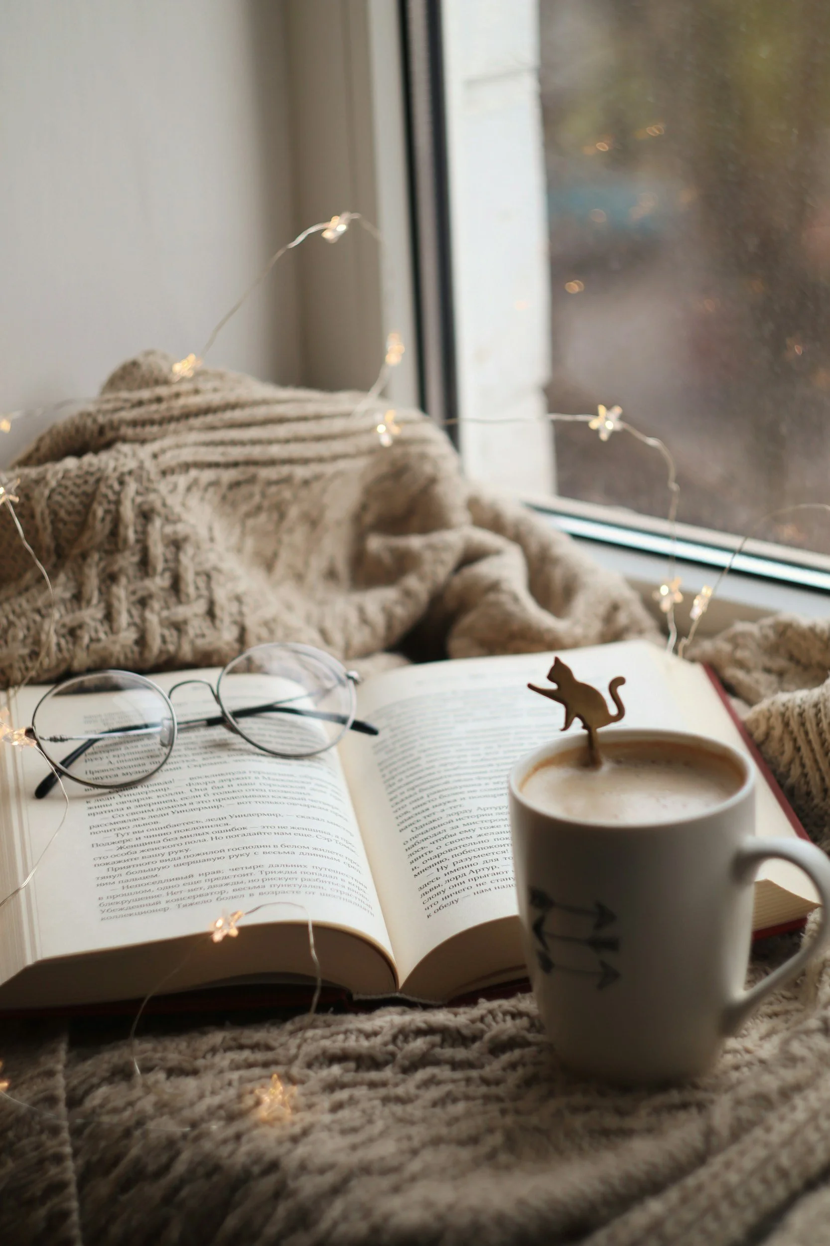 An open book with text in a foreign language, a pair of glasses, a cup of coffee with a small cat-shaped stirrer, a knitted blanket, a string of fairy lights, and a window with a blurred outdoor view.
