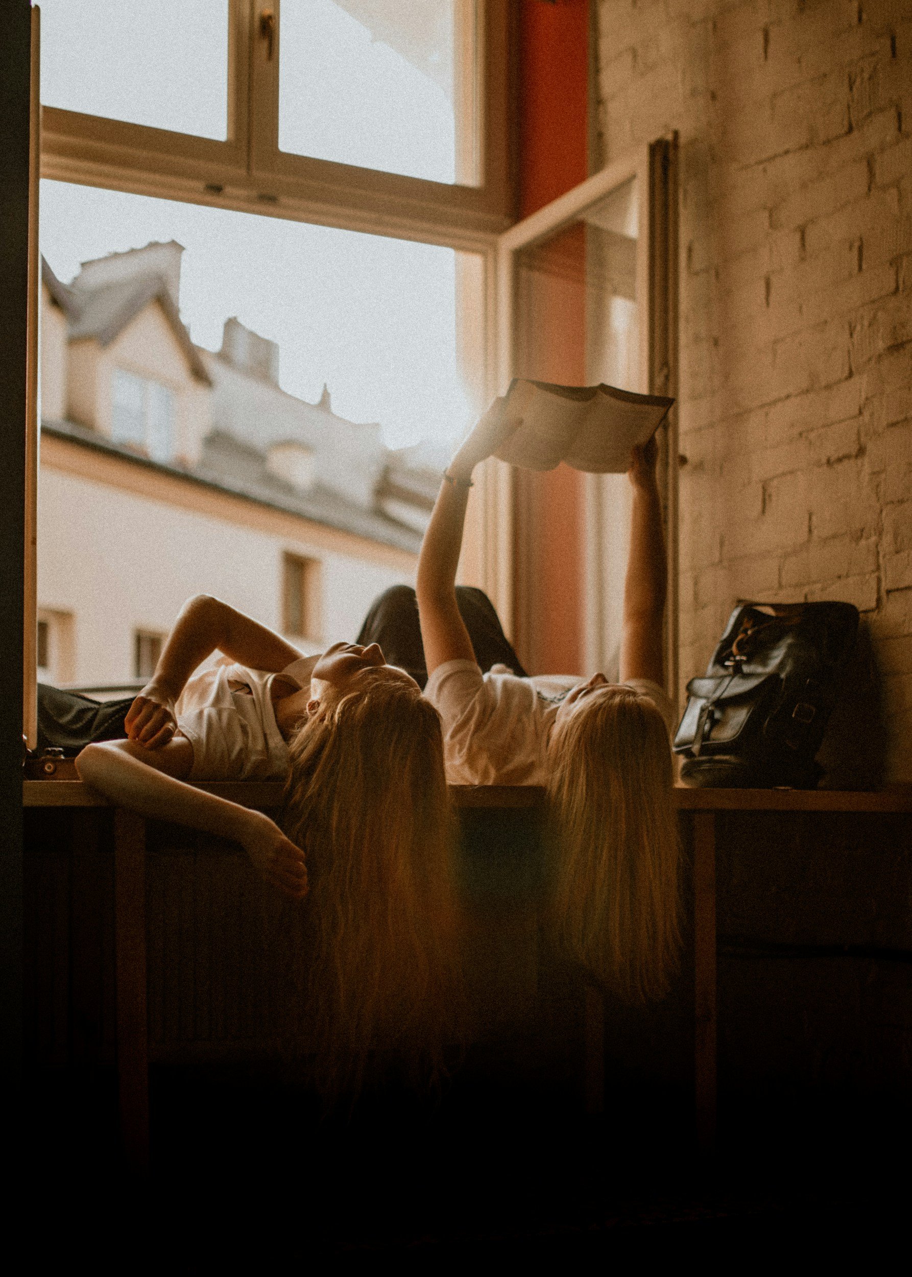 A person lying on a bed reading a book near a window with sunlight streaming in, with a brick wall on one side and a backpack on the bed.