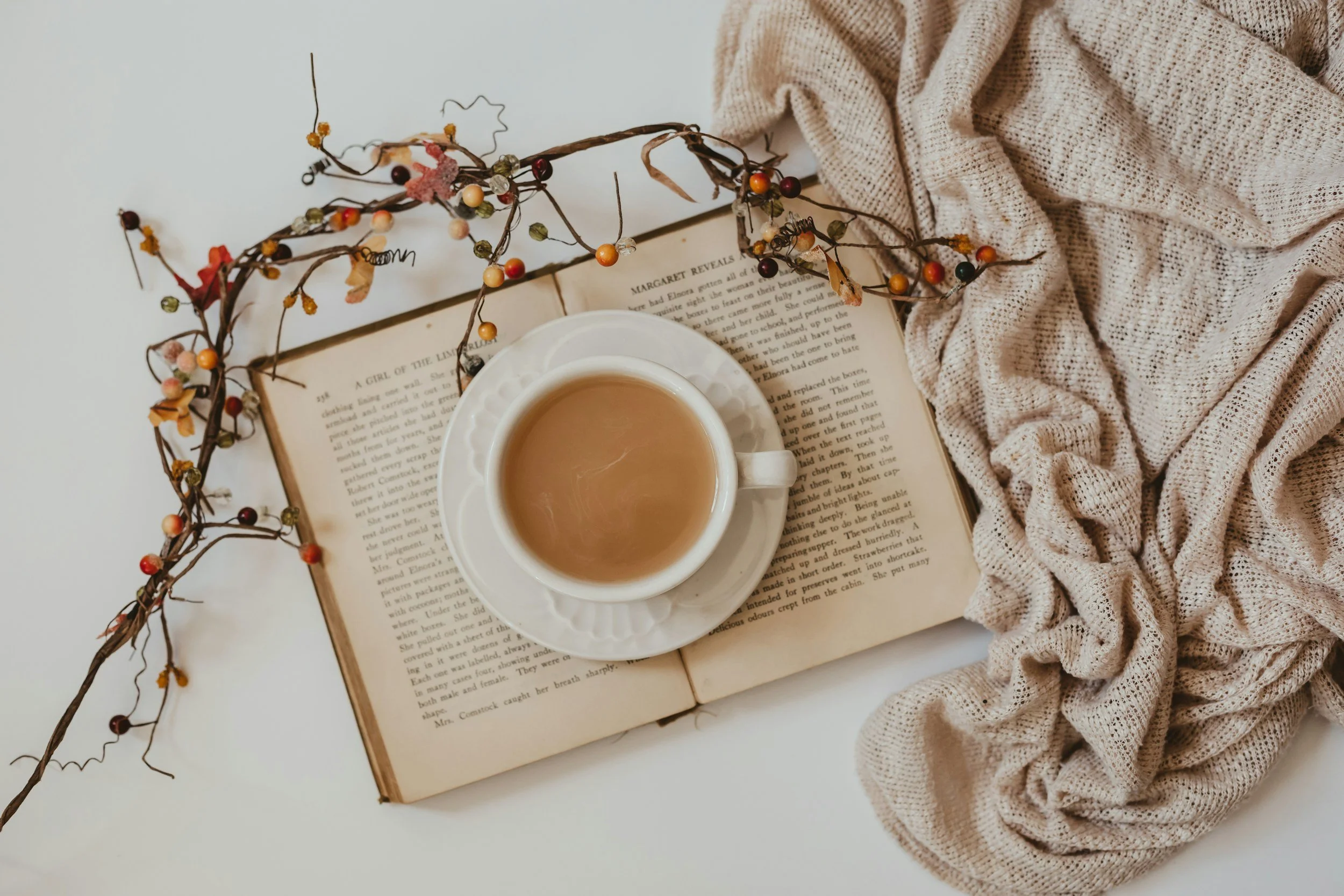 A cup of coffee on an open book, with a branch decorated with small colorful beads and dried leaves, and a beige knitted blanket.
