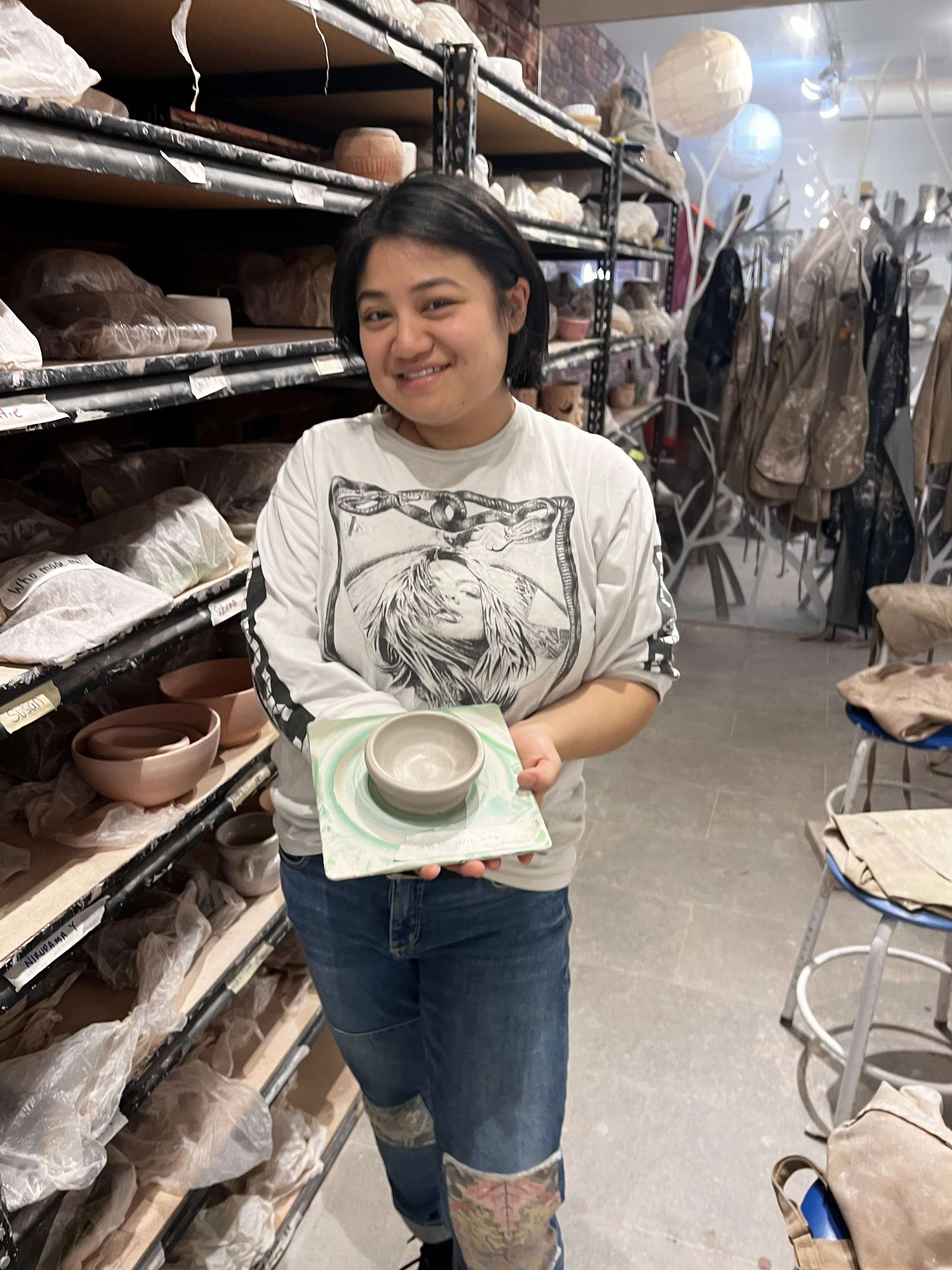 A young woman smiling and holding a ceramic bowl on a small ceramic plate in a pottery studio with shelves of ceramics and bags of clay in the background.