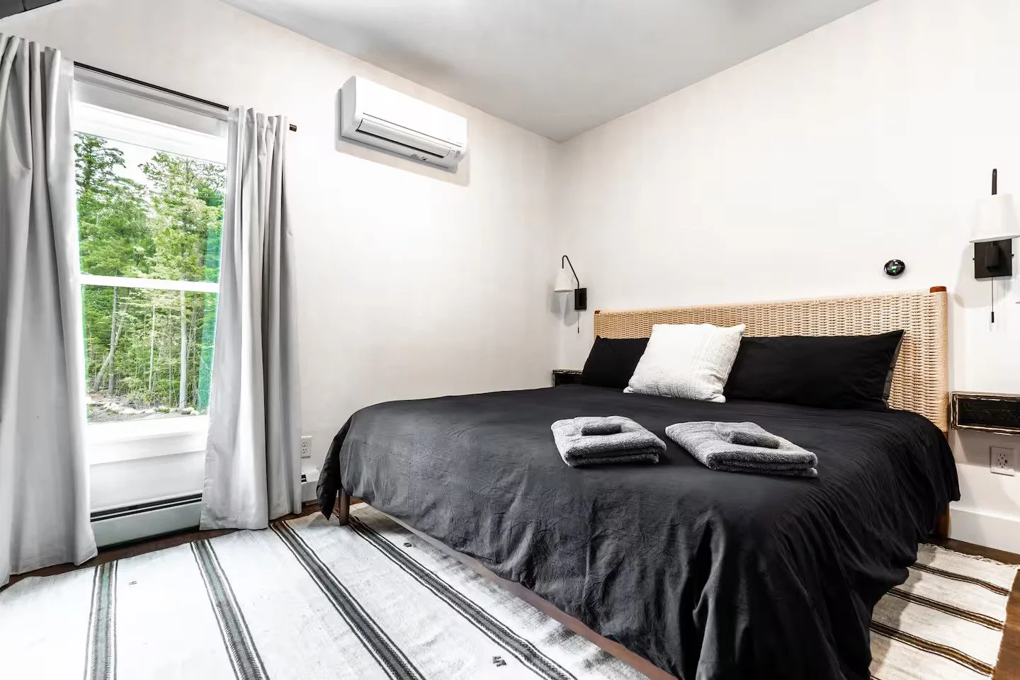 A bedroom with a large bed featuring a black bedspread, a white pillow, and two neatly folded gray towels. The room has white walls, a window with white curtains showing greenery outside, and a wall-mounted air conditioner above the window.