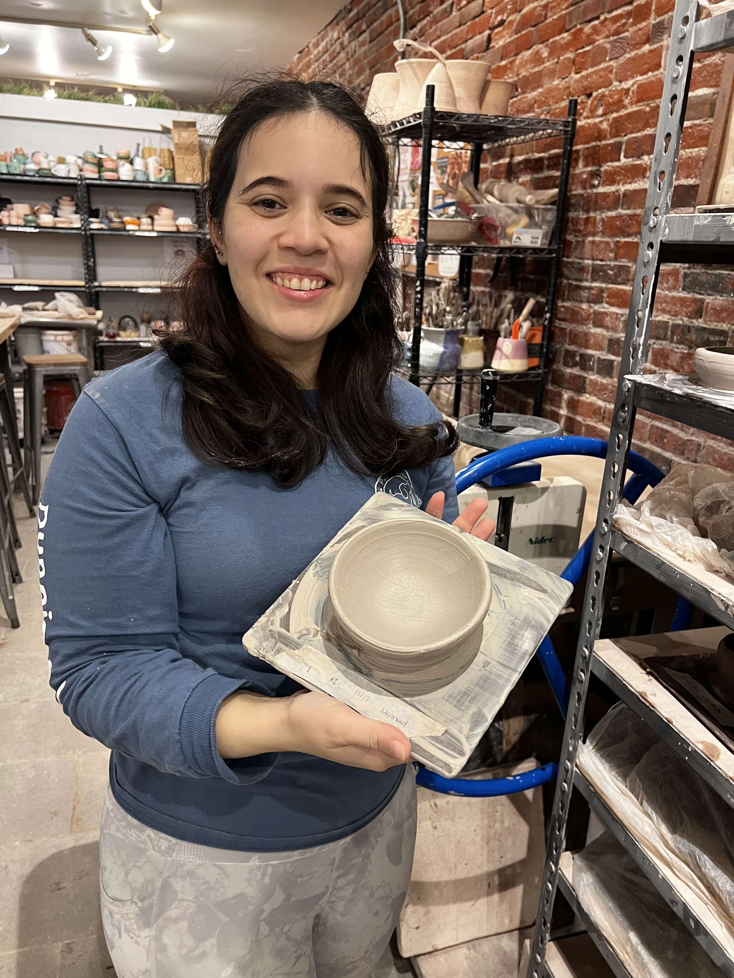 A woman holding a ceramic bowl in a pottery studio with shelves of pottery and tools in the background.