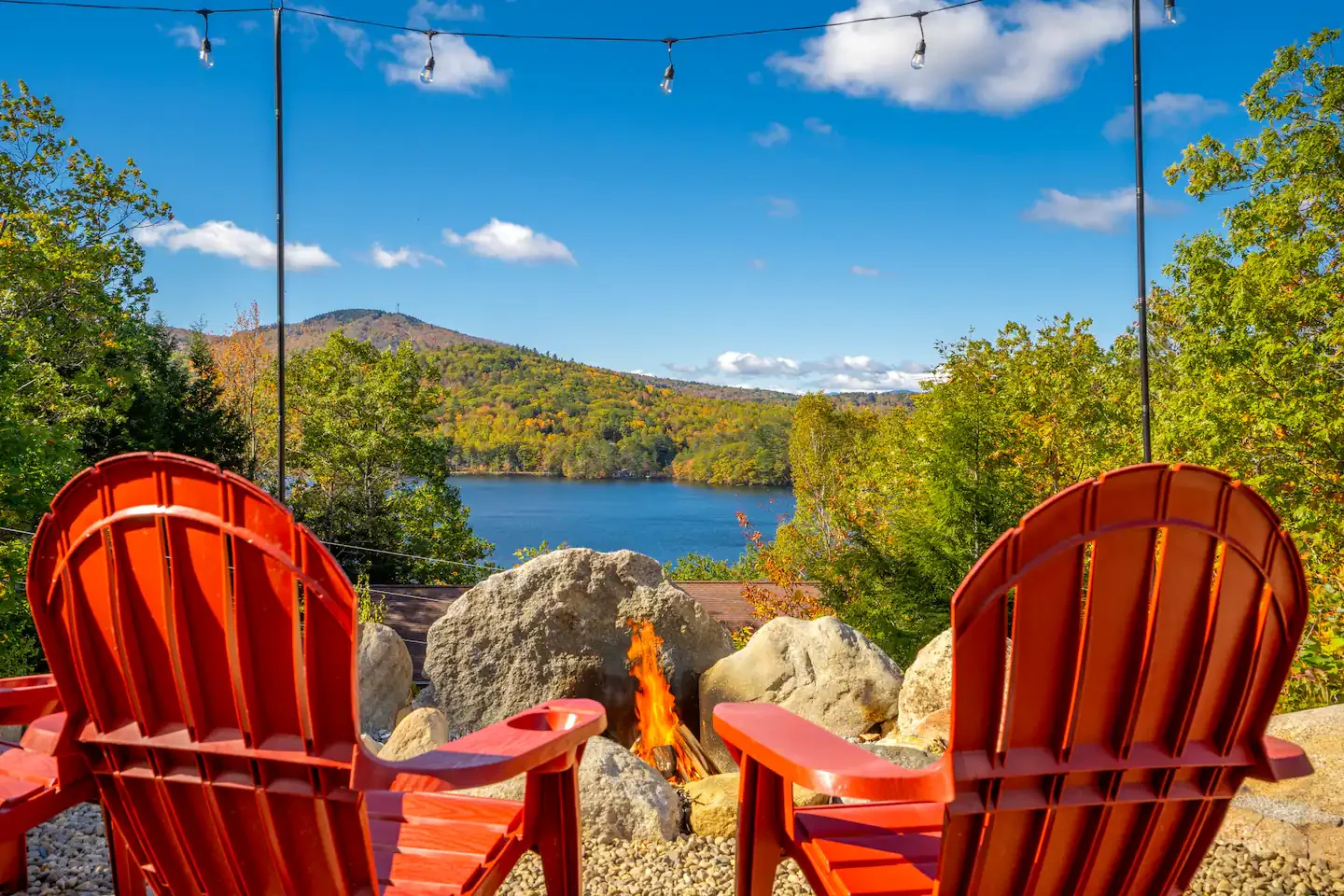 Two red Adirondack chairs facing a small campfire, overlooking a lake surrounded by green trees and hills under a blue sky with scattered clouds.