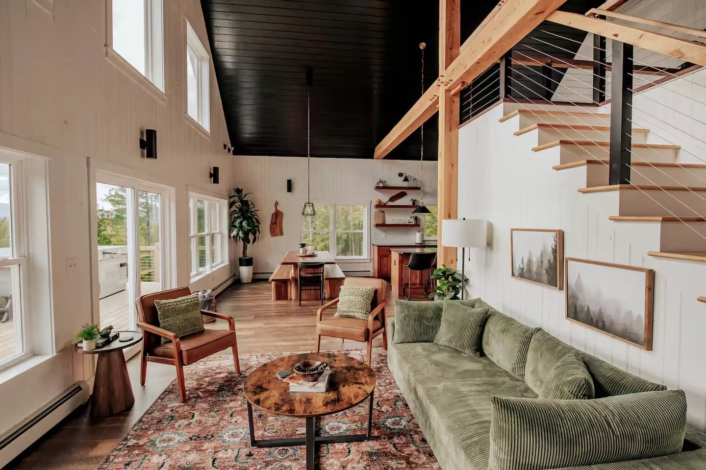A spacious living room with large windows, a green sofa, wooden chairs, a round coffee table, and a staircase with wooden steps and black metal railing, decorated with framed artwork and indoor plants.