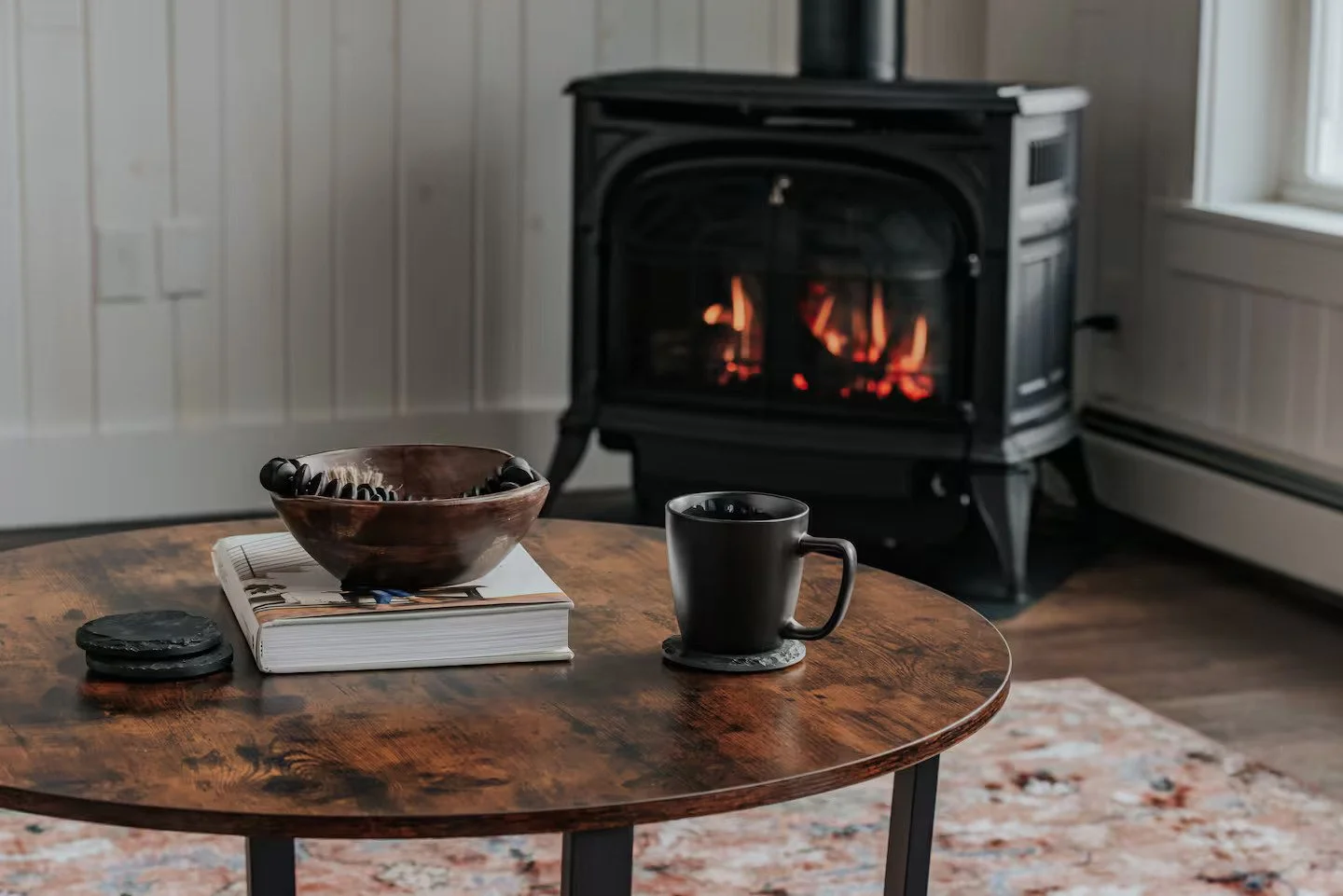 Living room with a round wooden table, a black mug on a coaster, an open book, a black rocky object, and a brown bowl with a black telephone inside it. A fireplace with a fire is in the background, next to a window.