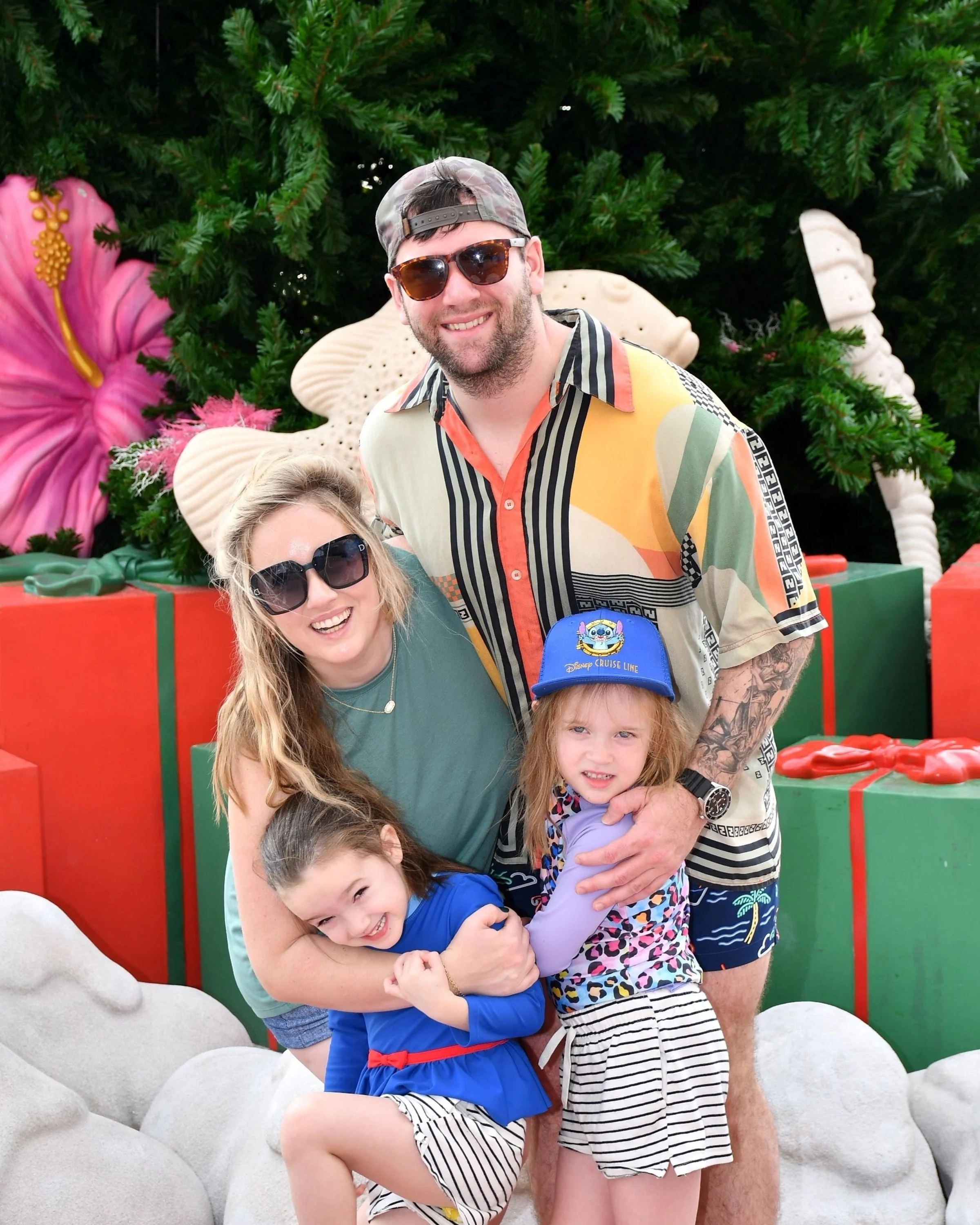 Group of people smiling and posing in front of a decorative outdoor setting with large flower and leaf sculptures, some wearing sunglasses and colorful casual clothing.