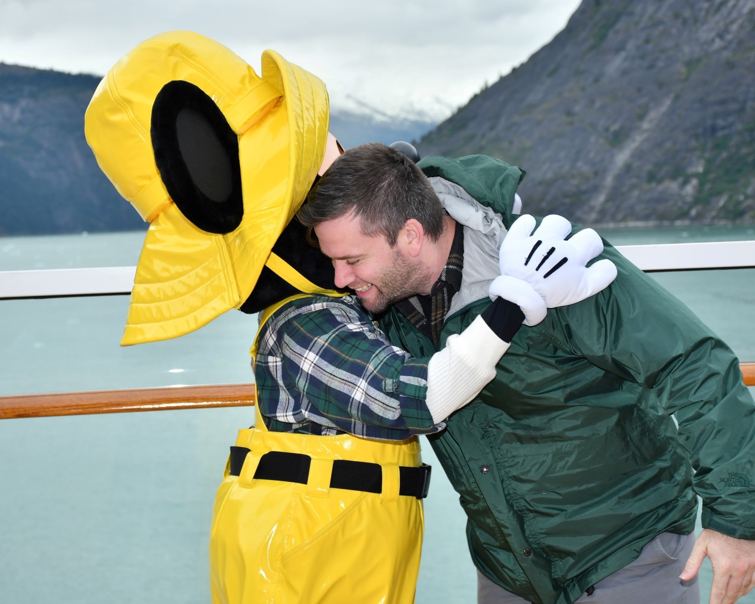 A man and a woman sharing a hug on a boat in a scenic outdoor location with mountains and water in the background. The woman is wearing a yellow jacket with a hood, and the man is wearing a green jacket with white gloves. They are smiling and appear happy.