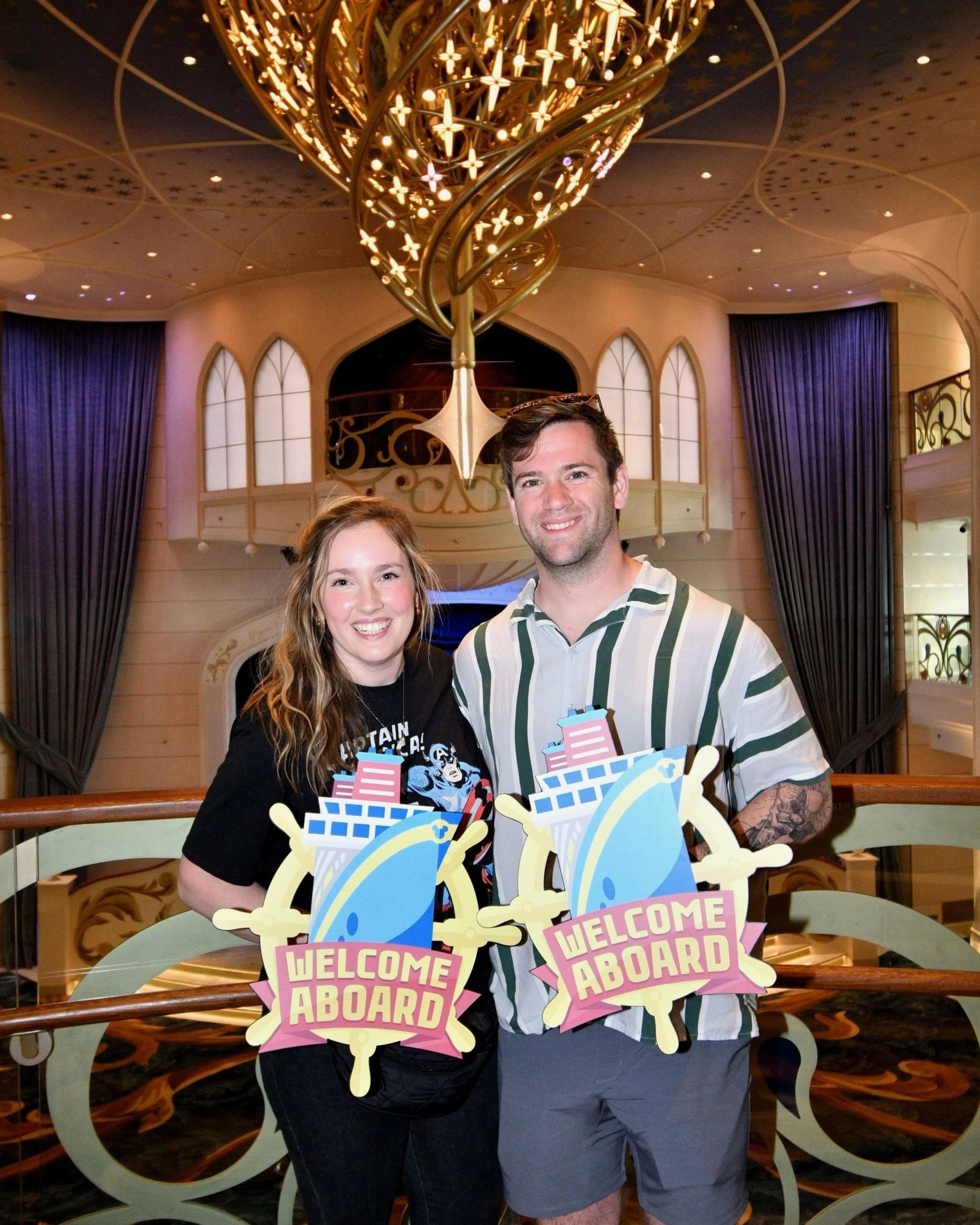 Two smiling young adults, a woman with wavy, shoulder-length red hair wearing a black T-shirt with Superman logo and text, and a man with short brown hair wearing a striped shirt, standing behind a decorative sign that says 'WELCOME ABOARD' in an indoor setting with ornate chandeliers and purple curtains in the background.