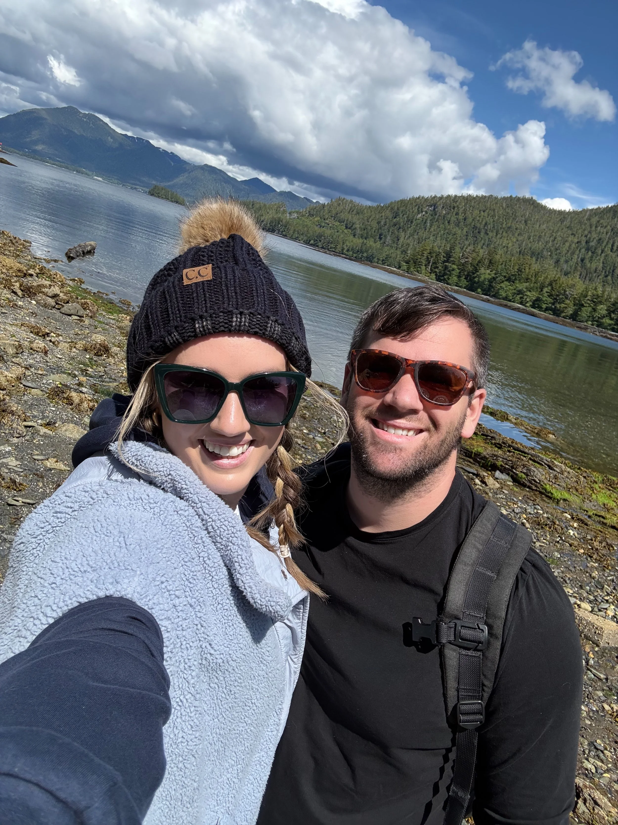 Smiling woman and man taking a selfie outdoors near a lake with mountains and trees in the background, under partly cloudy sky.