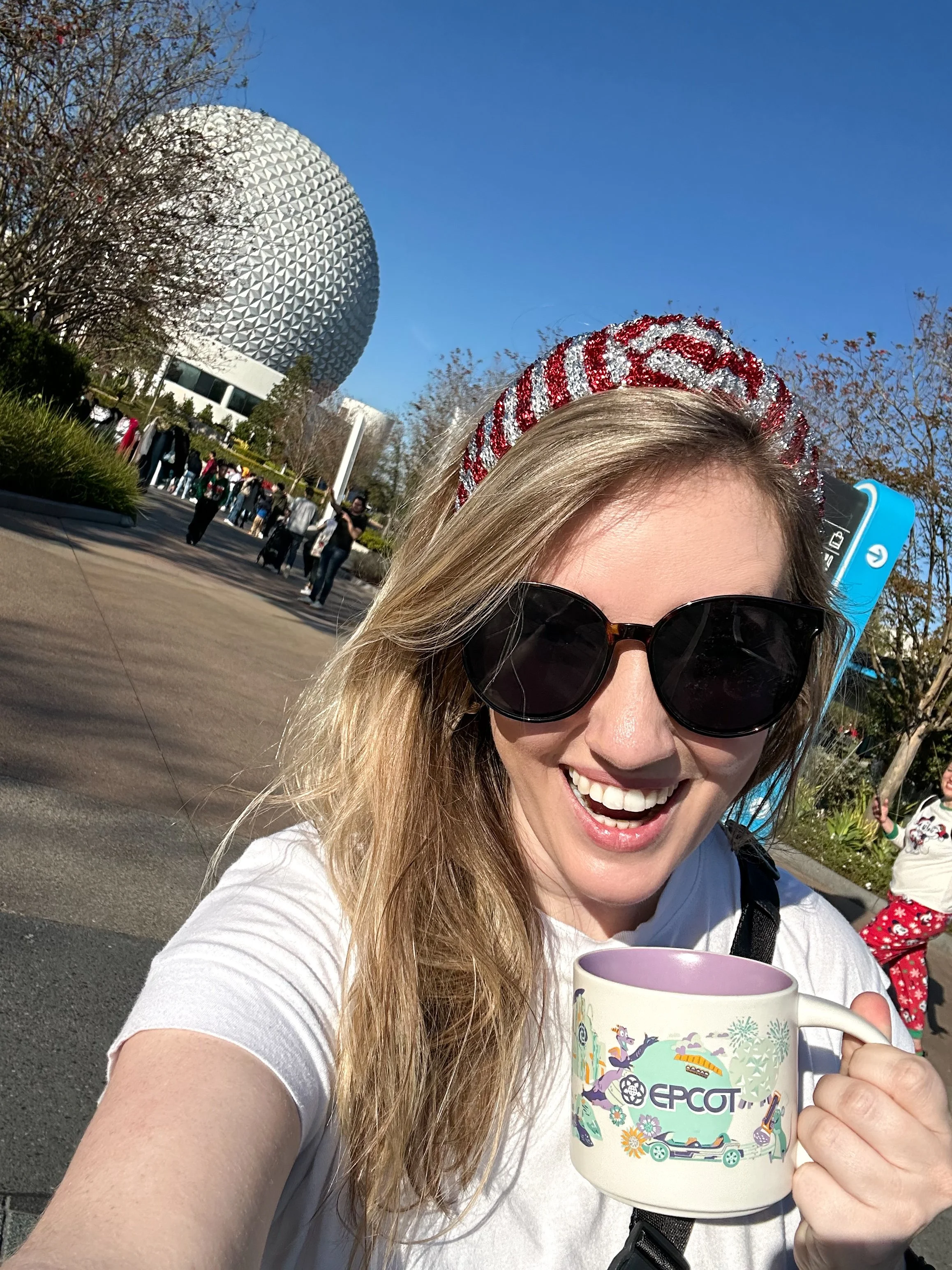 A woman with blonde hair, wearing sunglasses and a holiday headband, is smiling and holding a Disney-themed EPCOT mug in front of the EPCOT ball at Disney World, with other park visitors and trees in the background on a clear sunny day.
