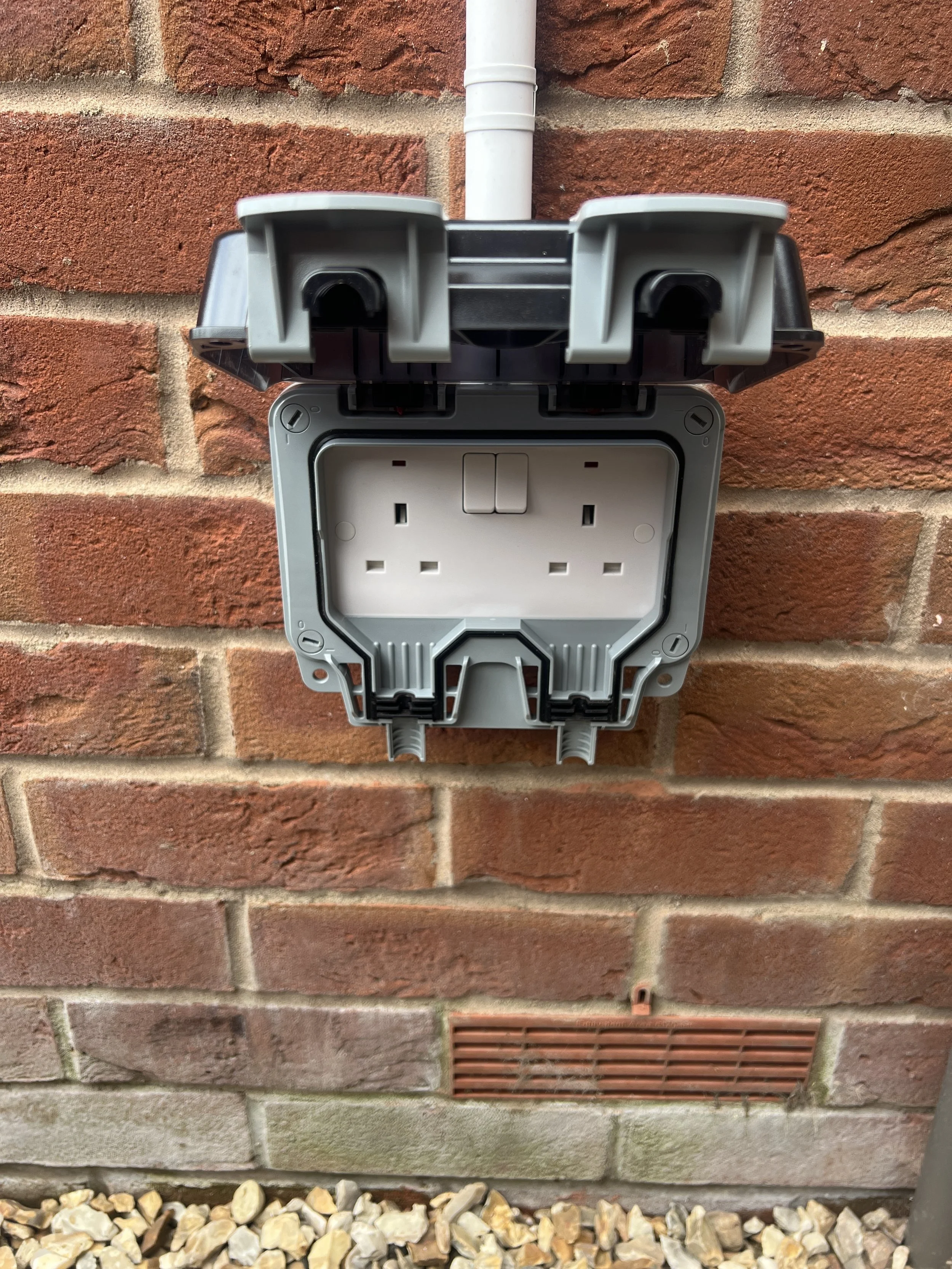 An empty outdoor electrical outlet box attached to a brick wall with a white conduit pipe at the top. The box is open and unpowered, with no cover or electrical connections visible.