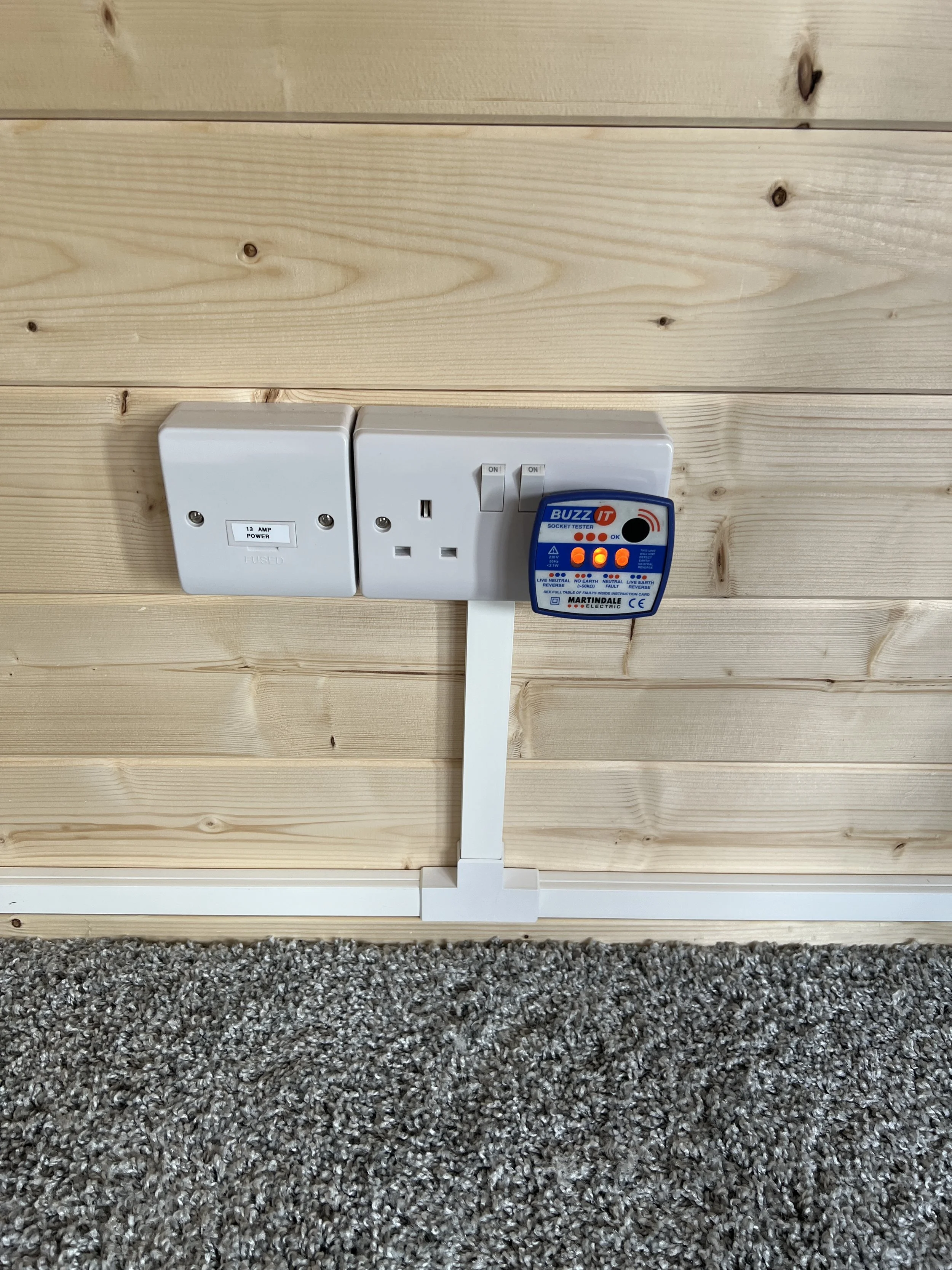 Electrical outlets and circuit tester mounted on a wooden wall with carpeted floor in front.