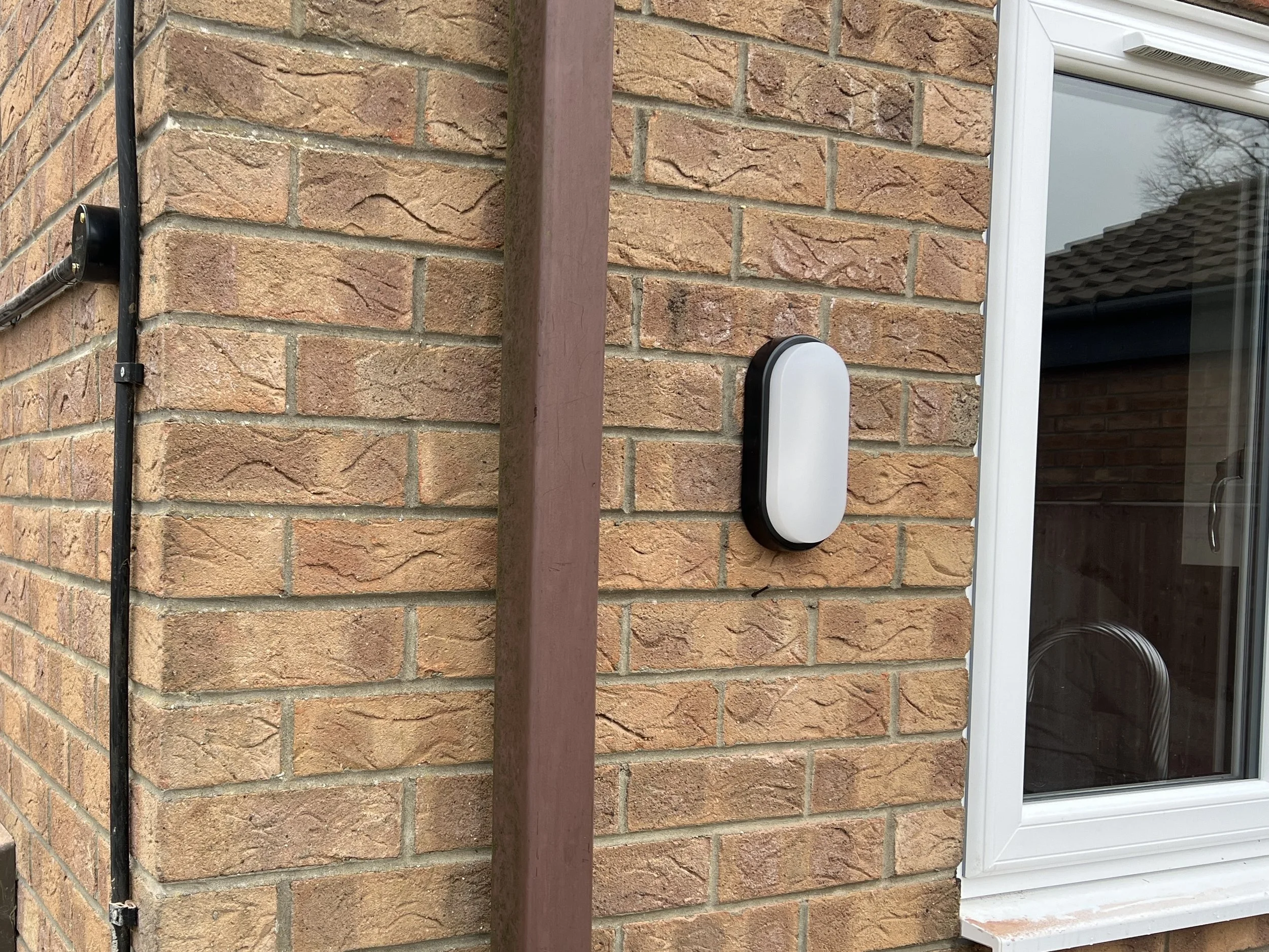 Close-up of a brick house wall with a white window and a round outdoor sensor or device mounted on the wall, with a wood post and an electrical conduit visible.