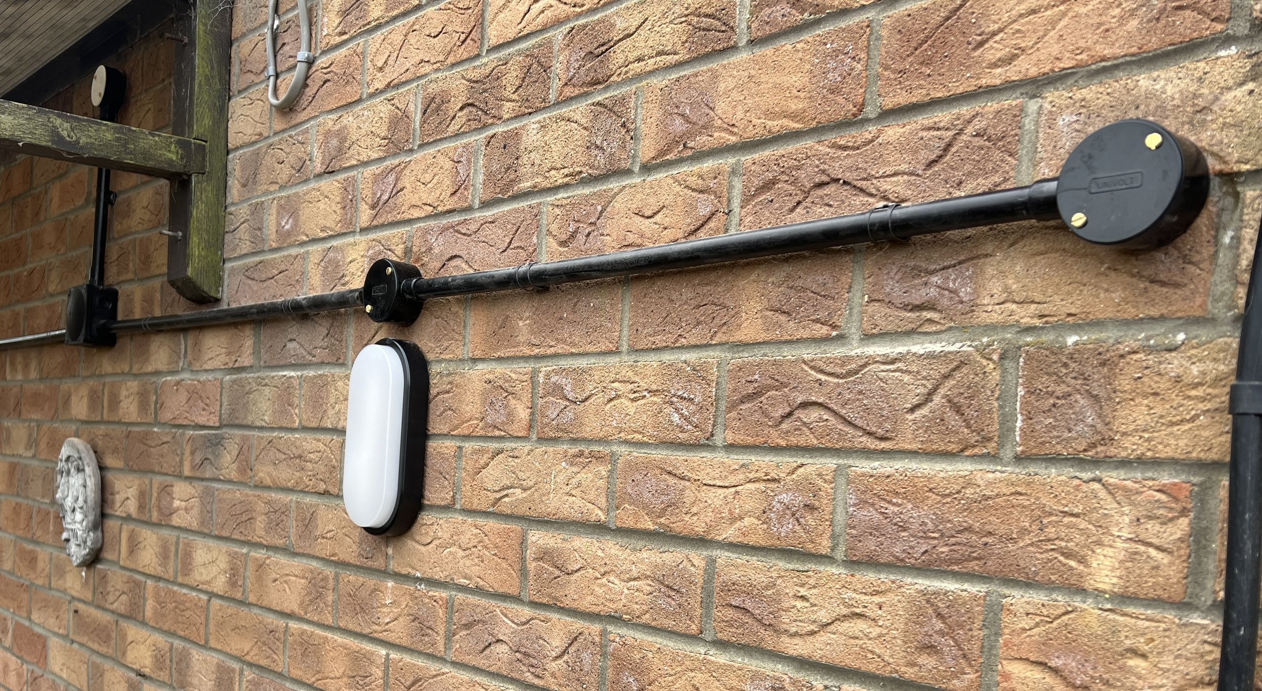 Close-up of a brick wall with electrical conduit, a black circular junction box, and a white outdoor light fixture mounted on it.