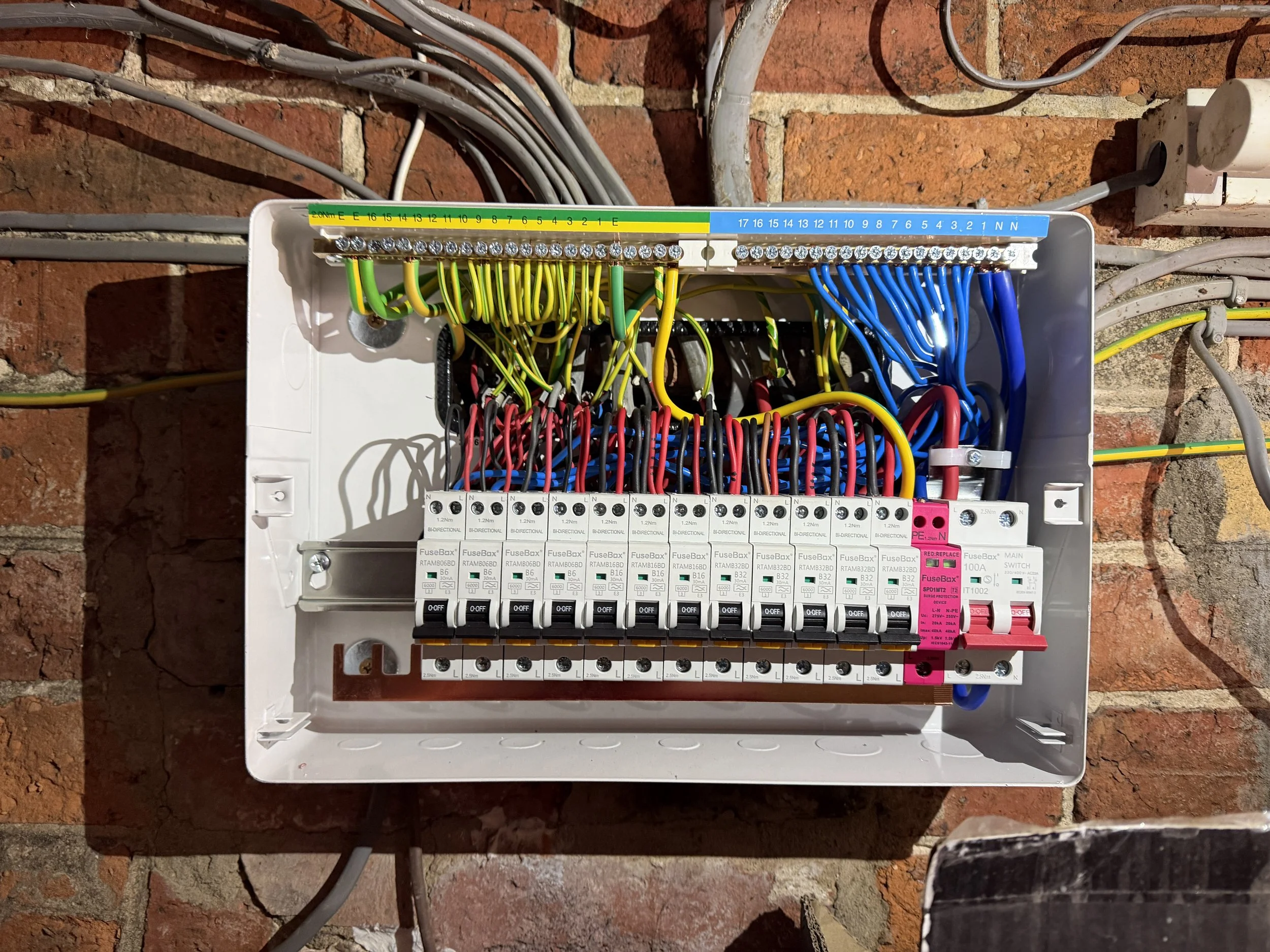 Electrical panel with multiple colored wires connected to a circuit breaker box mounted on a brick wall.