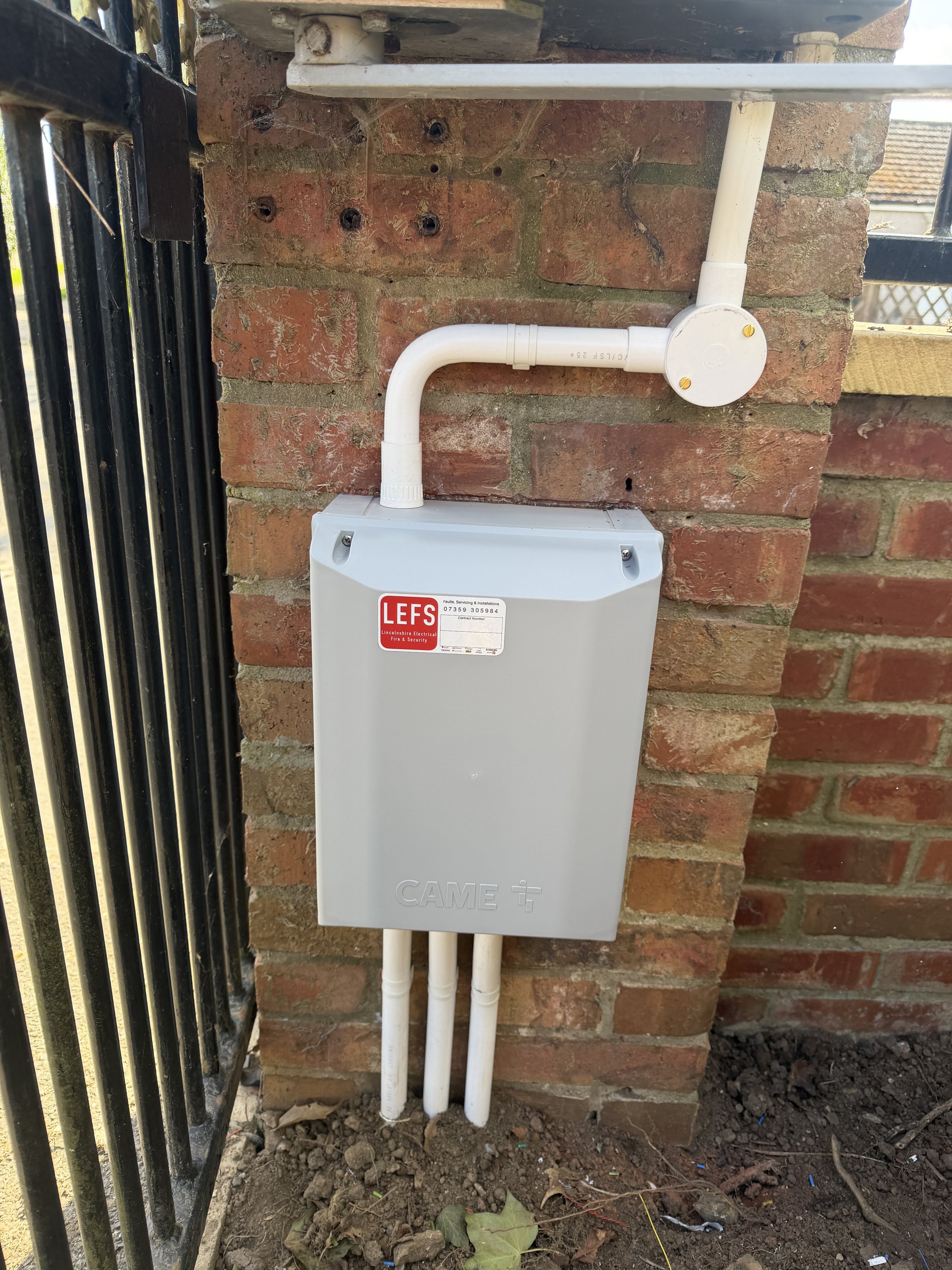 Electrical box mounted on a brick wall with white conduits connected to it, located outdoors near a black iron fence.