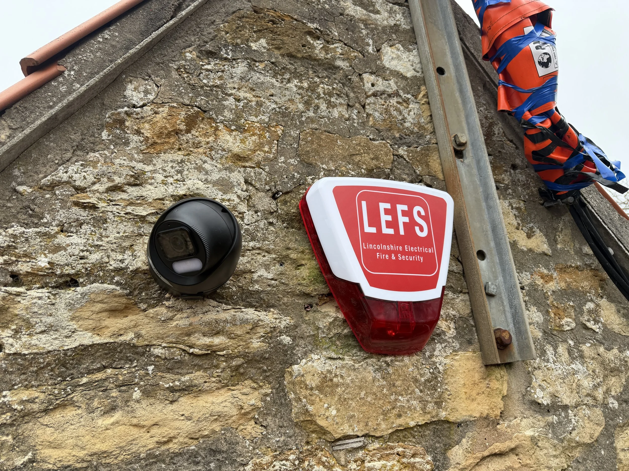Security camera and warning devices on a stone wall, including a red and white 'L.E.S.' fire and security sign, and bits of wiring tied with blue tape.