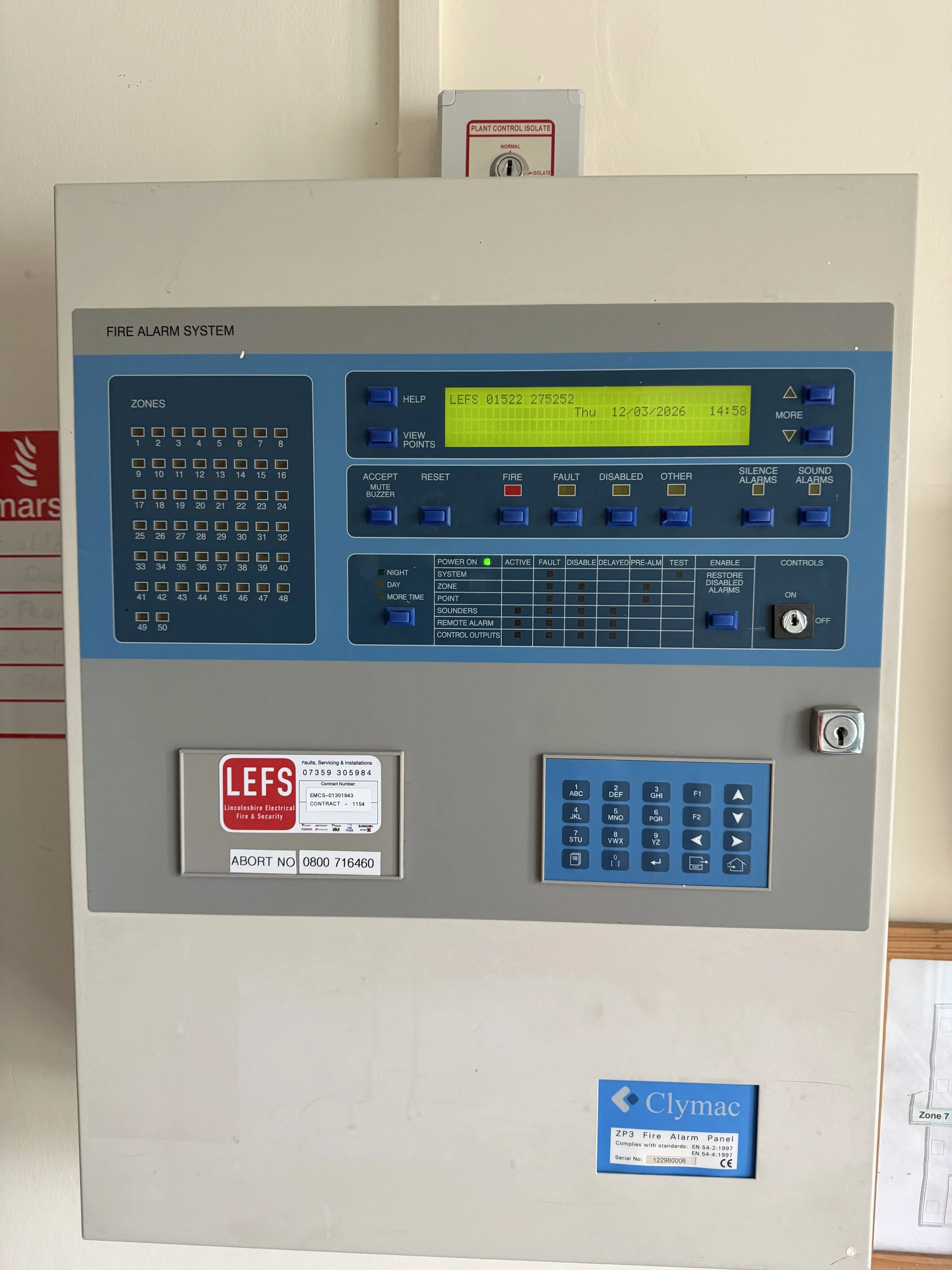 Electrical fire alarm control panel mounted on a wall with a blue and gray interface, a yellow digital screen displaying system information, and a lockable key switch at the bottom right corner.
