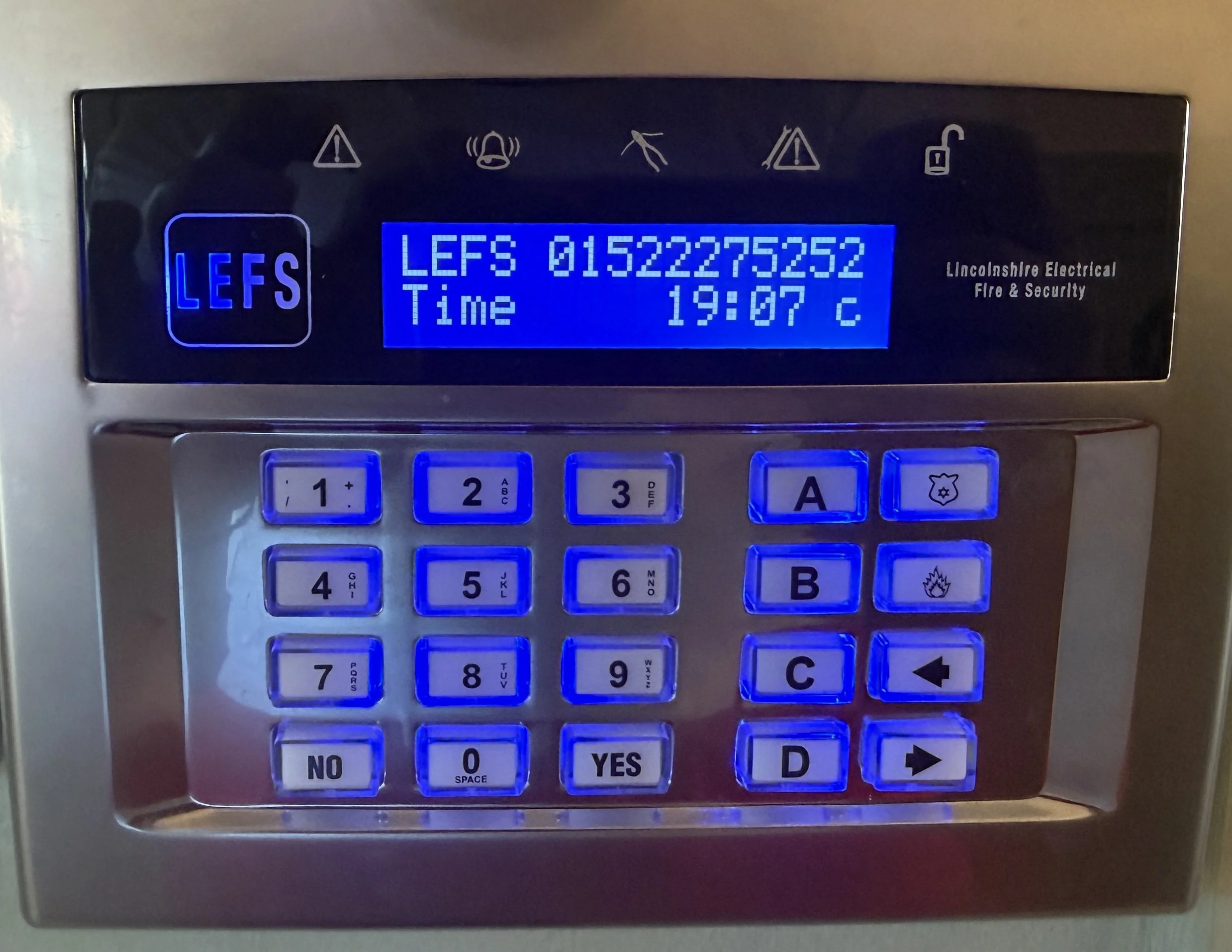 A digital wall-mounted fire alarm control panel with a blue backlit screen displaying the word "LEFS", a serial number, the time, and temperature. The panel has several illuminated blue buttons with labels including numbers, letters, and symbols. The surrounding area has several icons indicating various statuses and warnings.