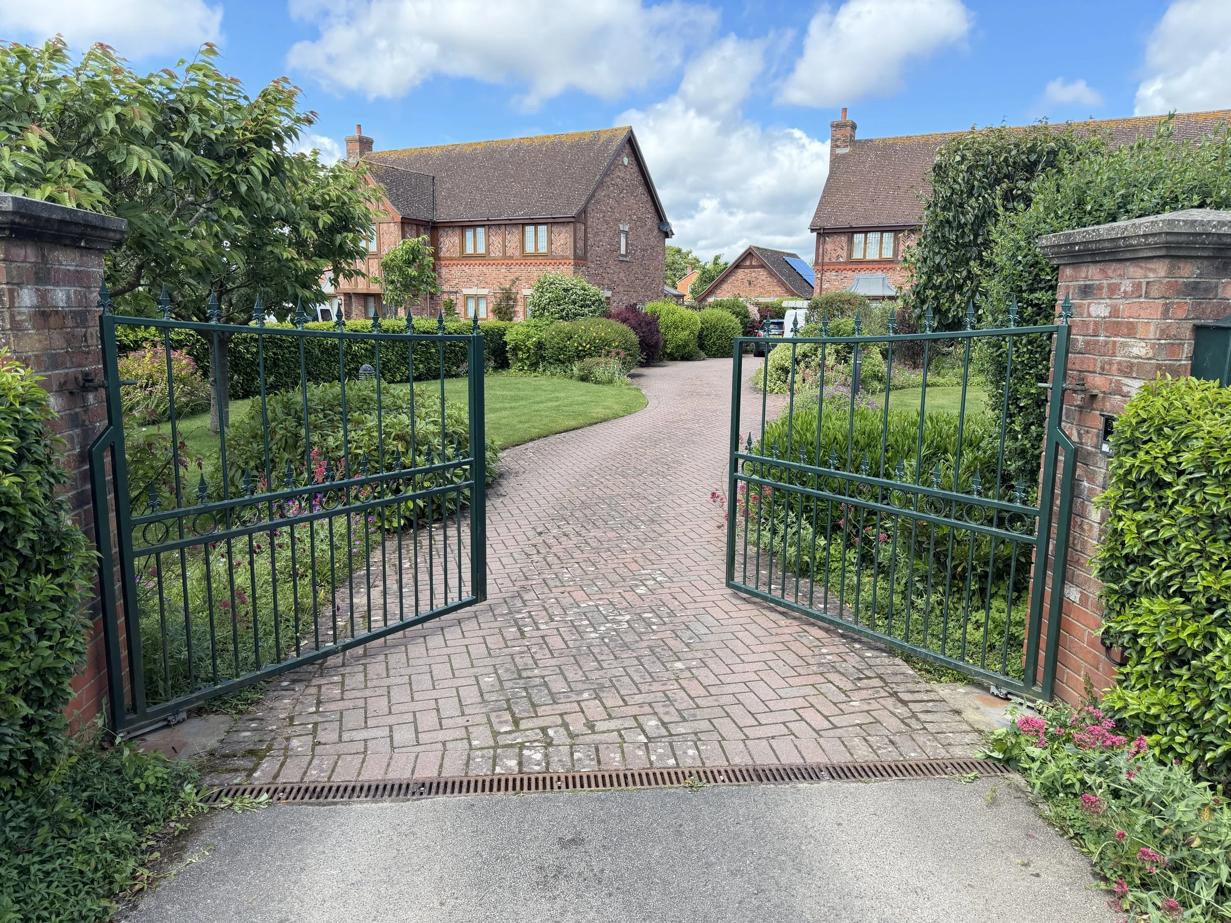A residential street view featuring brick houses with tiled roofs, a garden with bushes and plants, a brick pathway leading past a green metal gate, and a blue sky with some fluffy clouds.