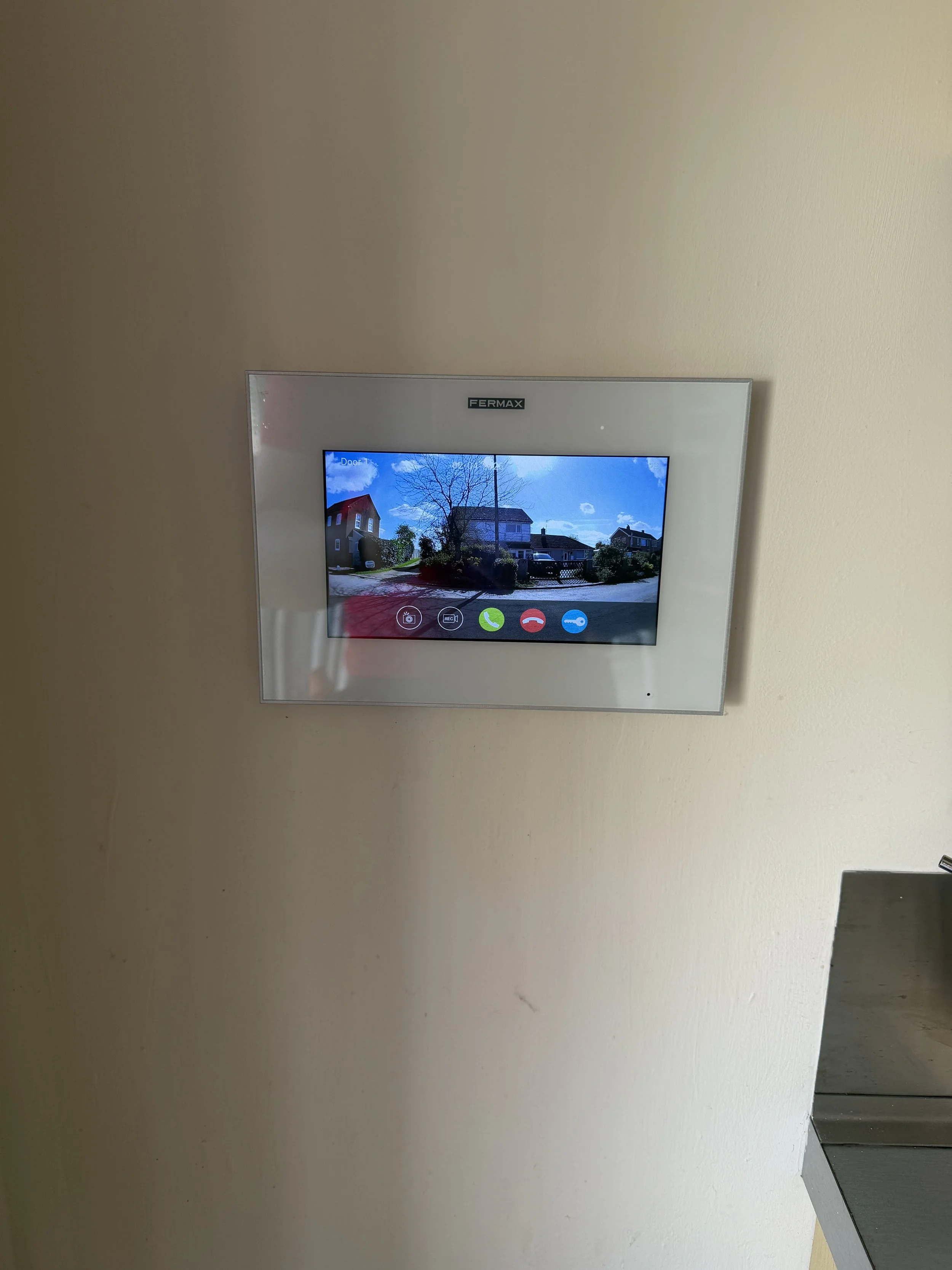 A beige wall with a mounted touch screen device showing a street scene with houses, a tree, and a clear blue sky.