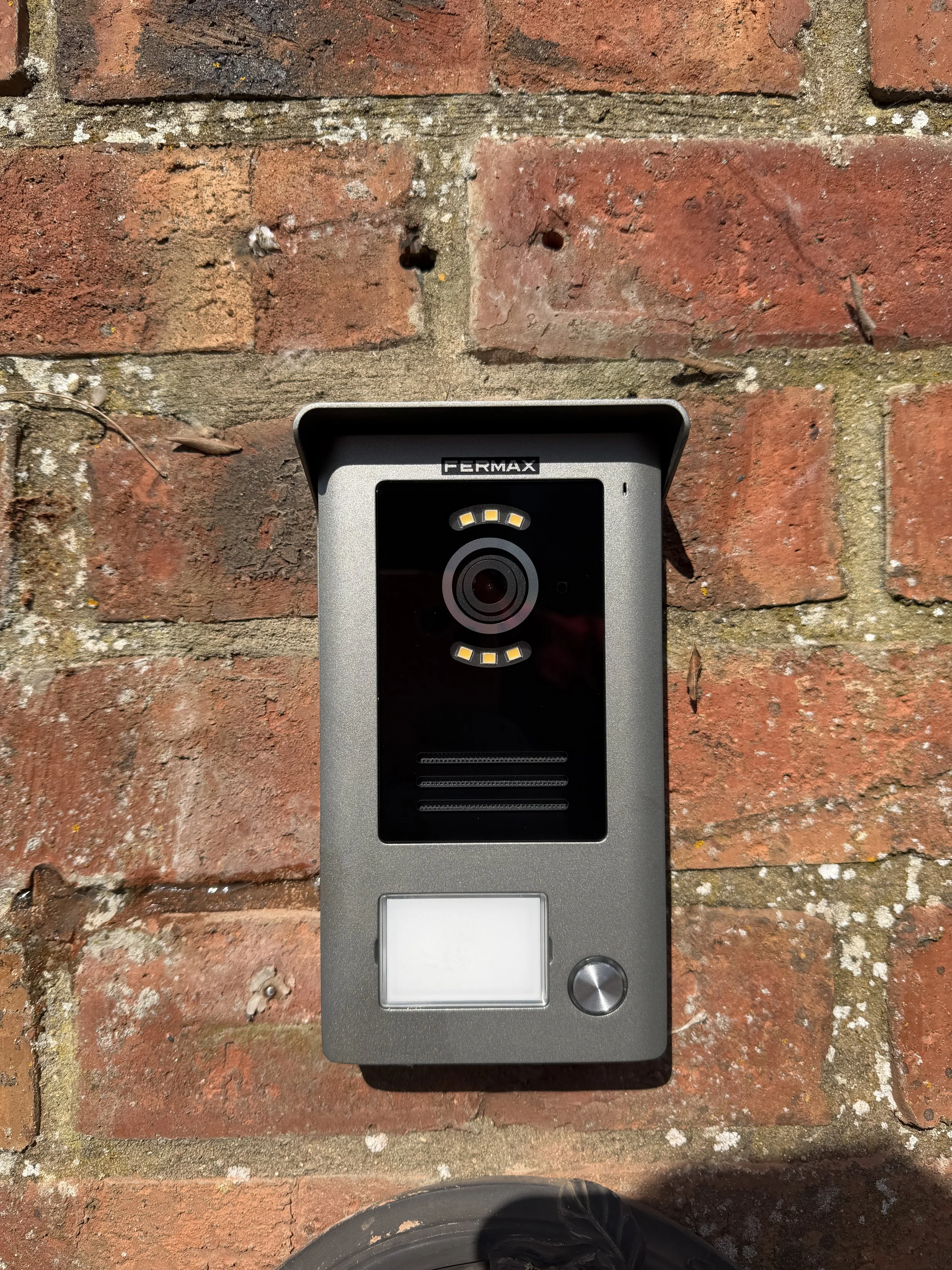 A modern doorbell and intercom system mounted on a red brick wall, with a plastic or metal casing, a camera at the top, a small screen or panel below, and a round button in the lower right corner.