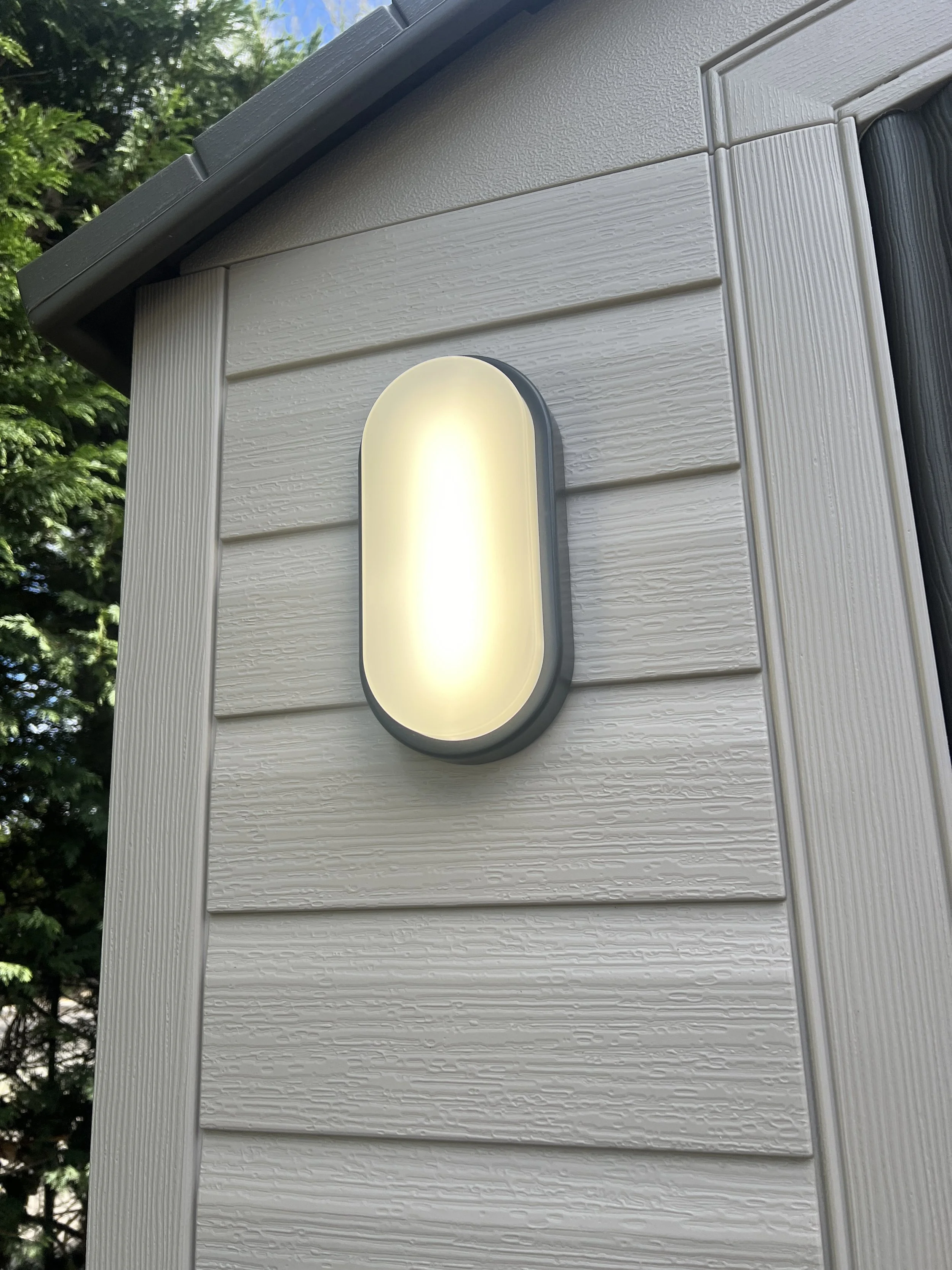 Close-up of a modern outdoor wall-mounted light fixture attached to a white textured siding house, with trees and blue sky in the background.