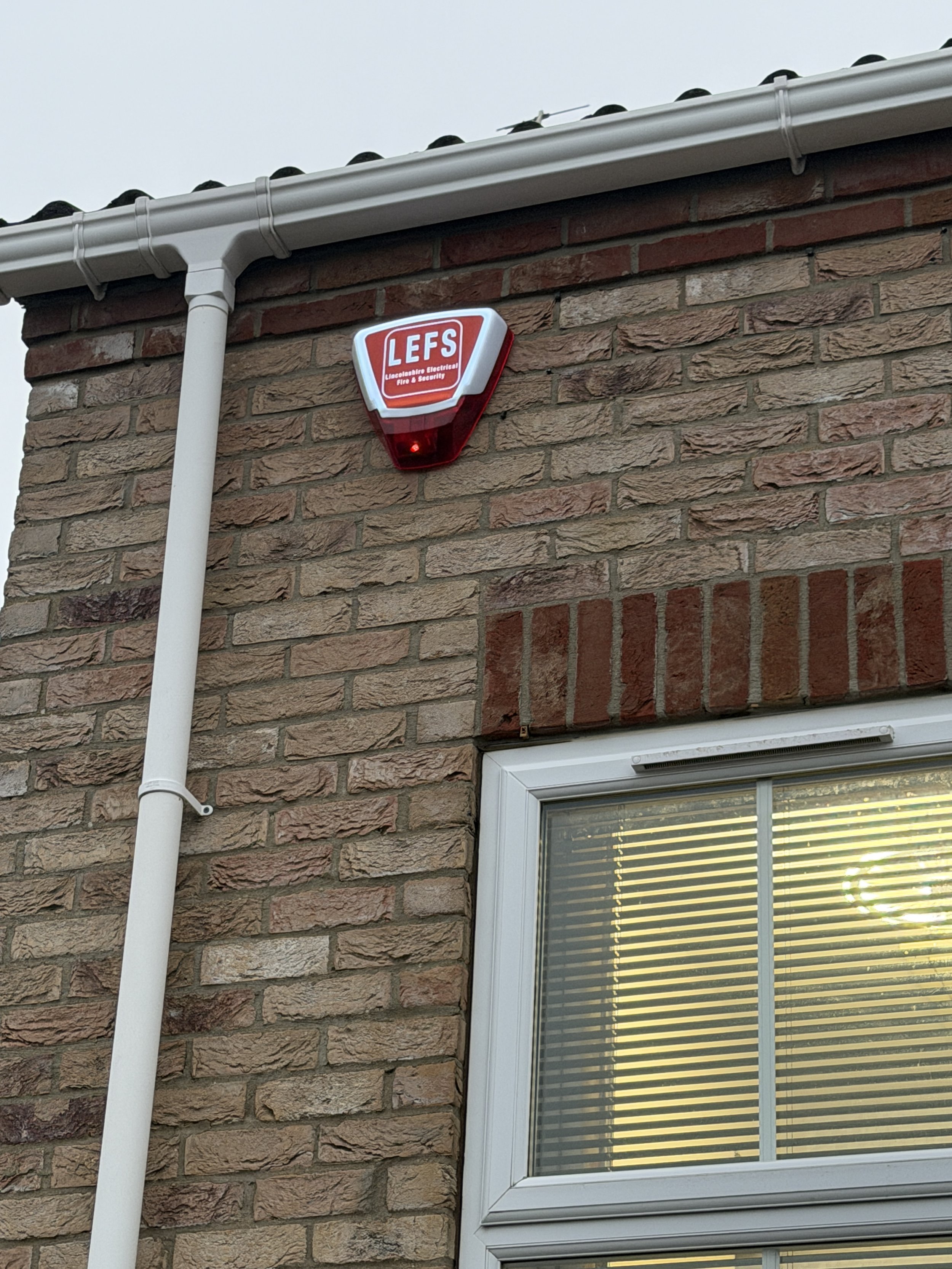 The image shows the exterior of a brick house with a window and a security alarm device on the wall. The alarm reads LEF, Lincolnshire Electrical Fire & Security.