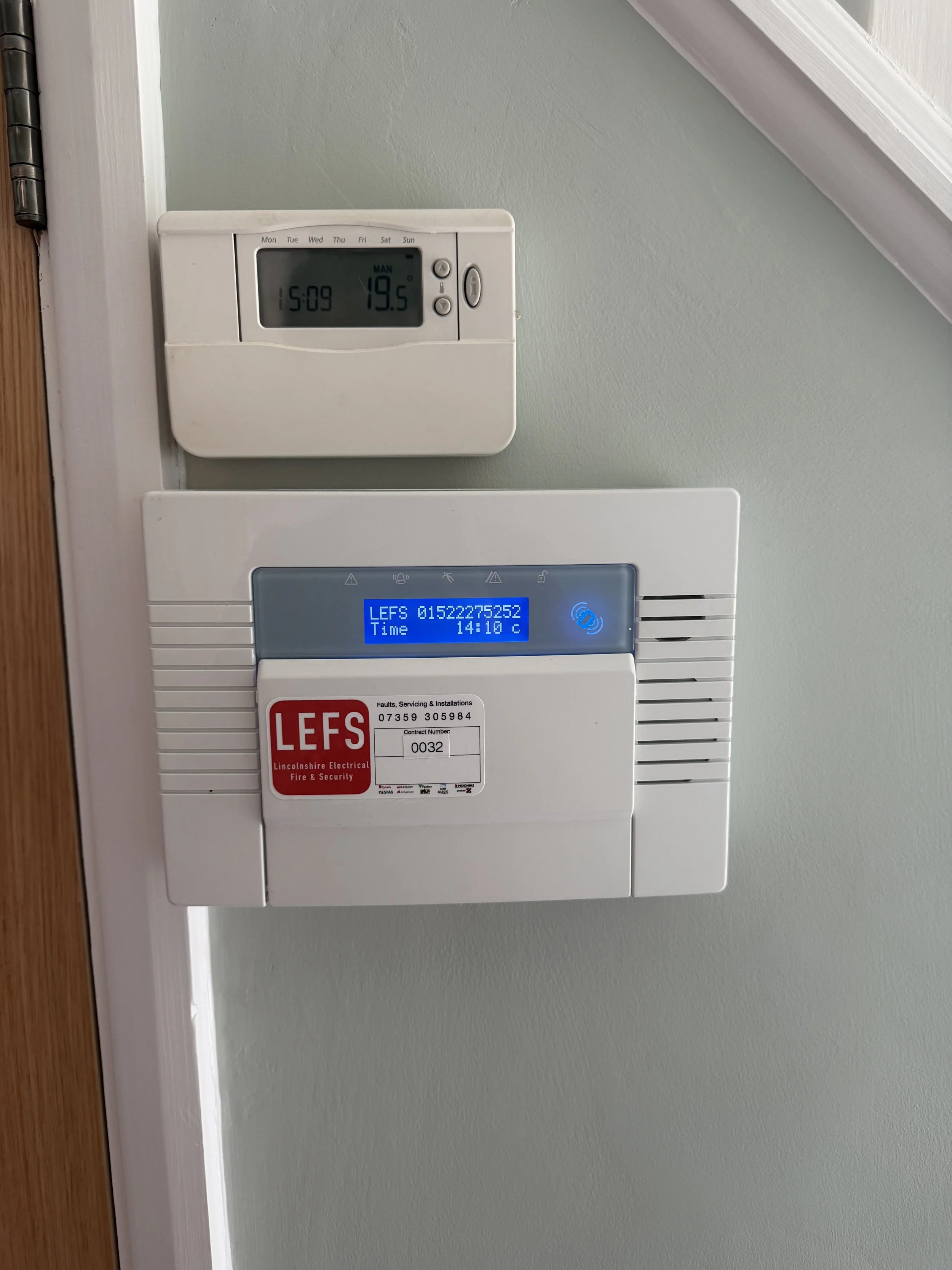 A digital thermostat displays the time as 15:09, the temperature as 19.5 degrees Celsius, and the day as Monday. Below, a white fire alarm system panel shows the LEFS logo and contact information.