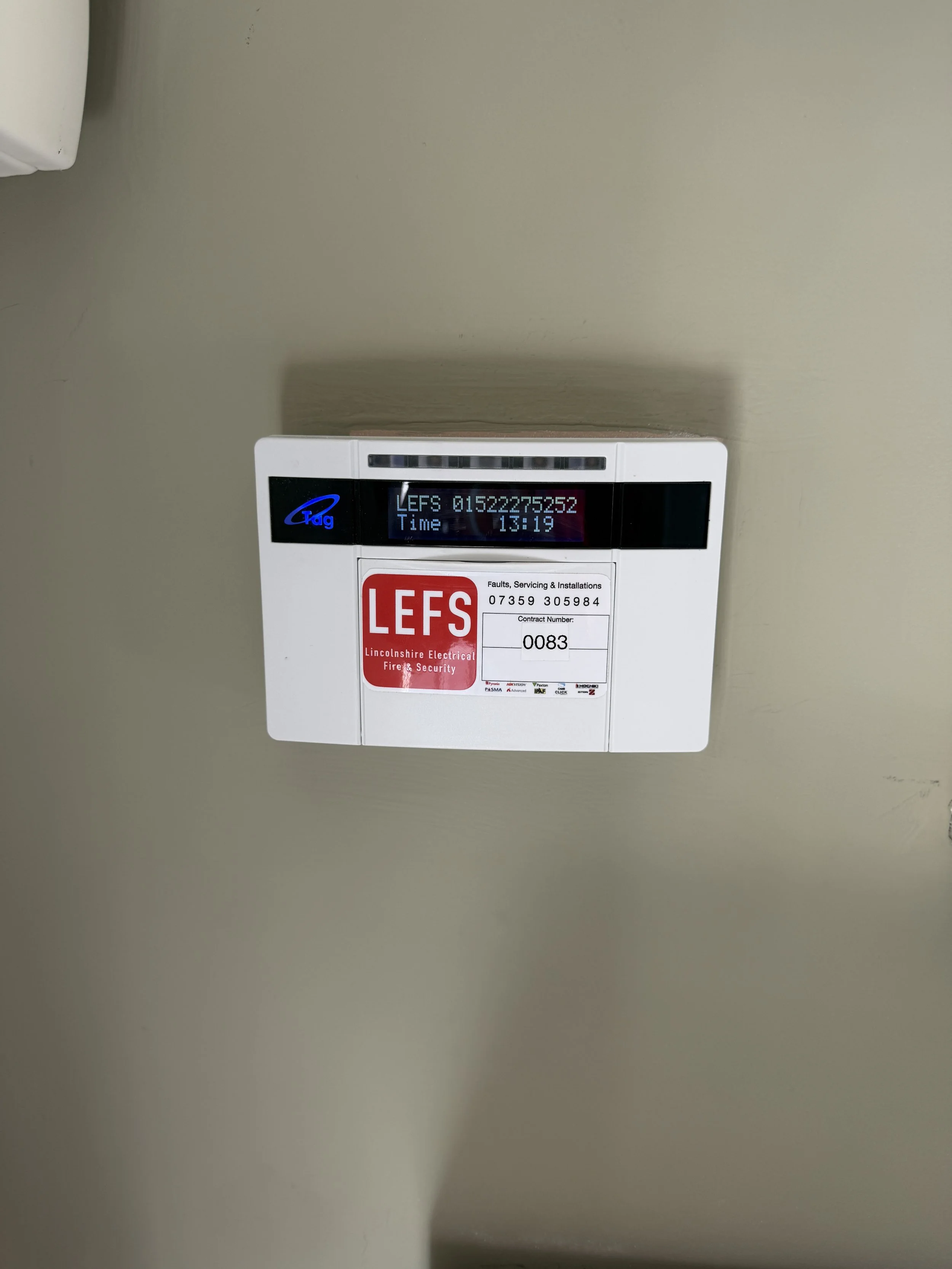 White electrical control box on a beige wall with a digital display showing a code, a time of 13:19, and a red sticker that reads 'LEFS' and 'Lincolnshire Electrical Fire & Security'.