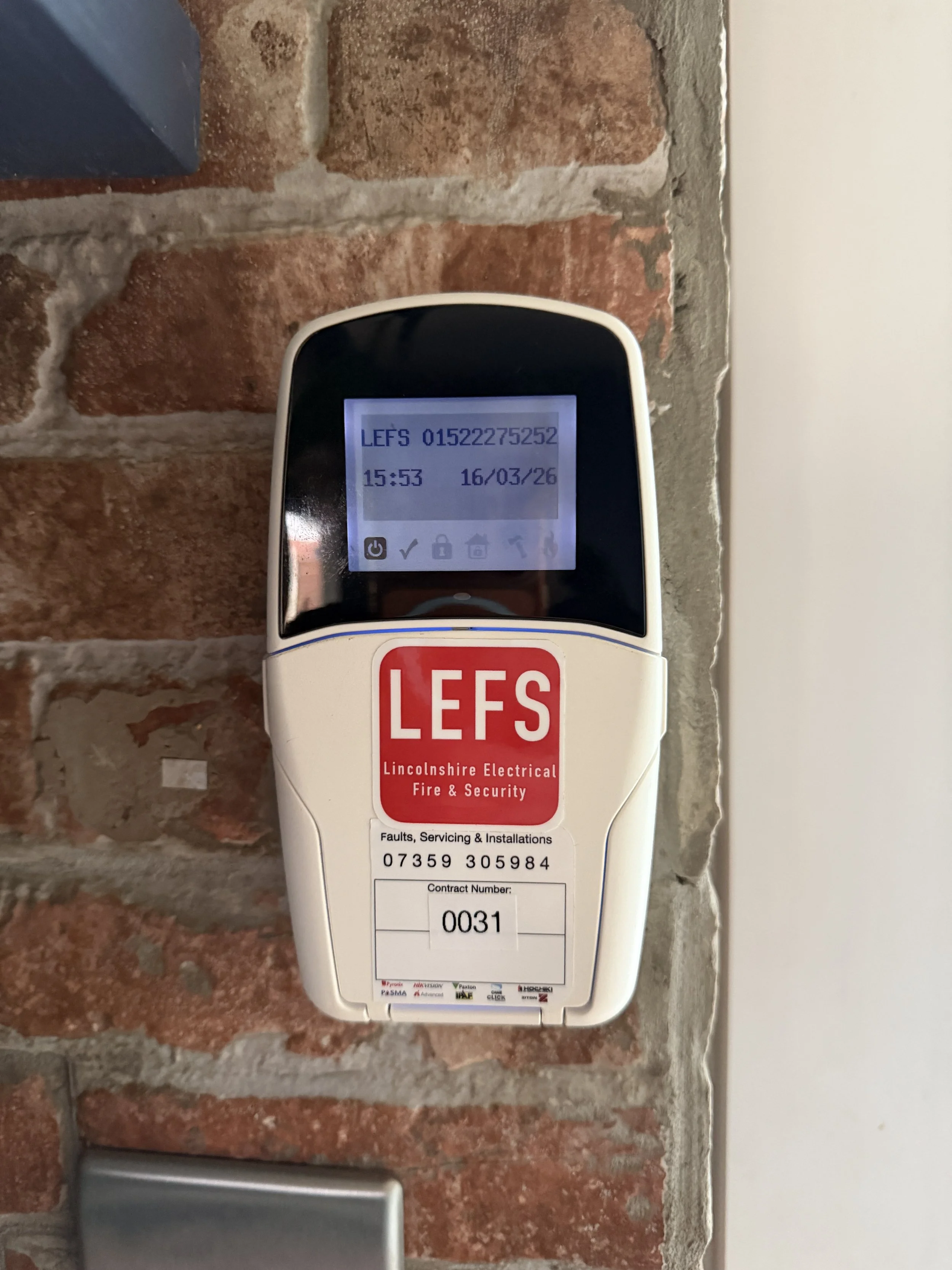 A fire alarm control panel mounted on a brick wall with an LCD screen displaying unit identification information, date, and time. The panel has a red and white sticker labeled "LEFS" from Lincolnshire Electrical Fire & Security.