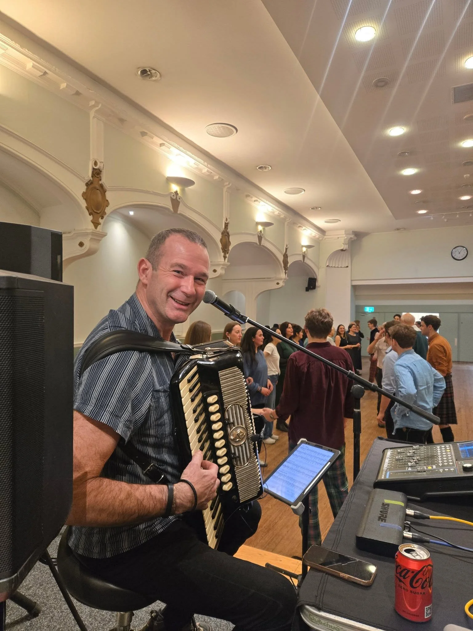 A man playing an accordion and smiling at a social gathering indoors, with people socializing in the background, a microphone, audio equipment, and a Coca-Cola can on the table. Clappy Doo Ceilidh band Scottish Ceilidh band Scotland music
