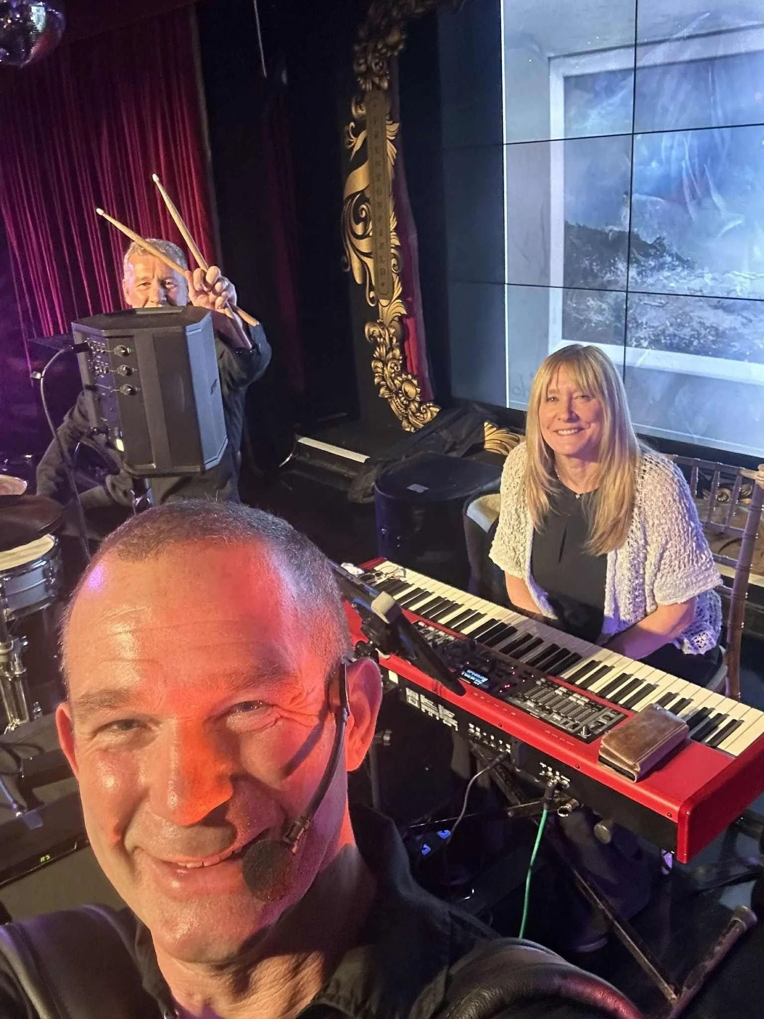 A man smiling at the camera wearing glasses, with a keyboard and electronic music equipment in front of him, and a woman sitting behind a keyboard, smiling. There are musical instruments and a large screen in the background.  Clappy Doo Ceilidh band 
