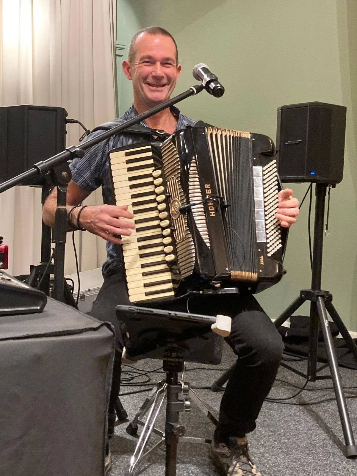 A man smiling while playing an accordion at a live music event with microphones and speakers around him.  Clappy Doo Ceilidh band Scottish Ceilidh band traditional Scottish music