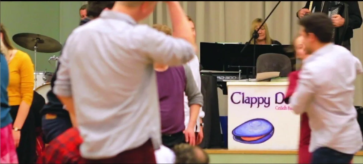People dancing at an event with a band playing on stage, including a pianist and a harmonica player, and a sign that reads "Clappy D" with a cartoon clam shell.