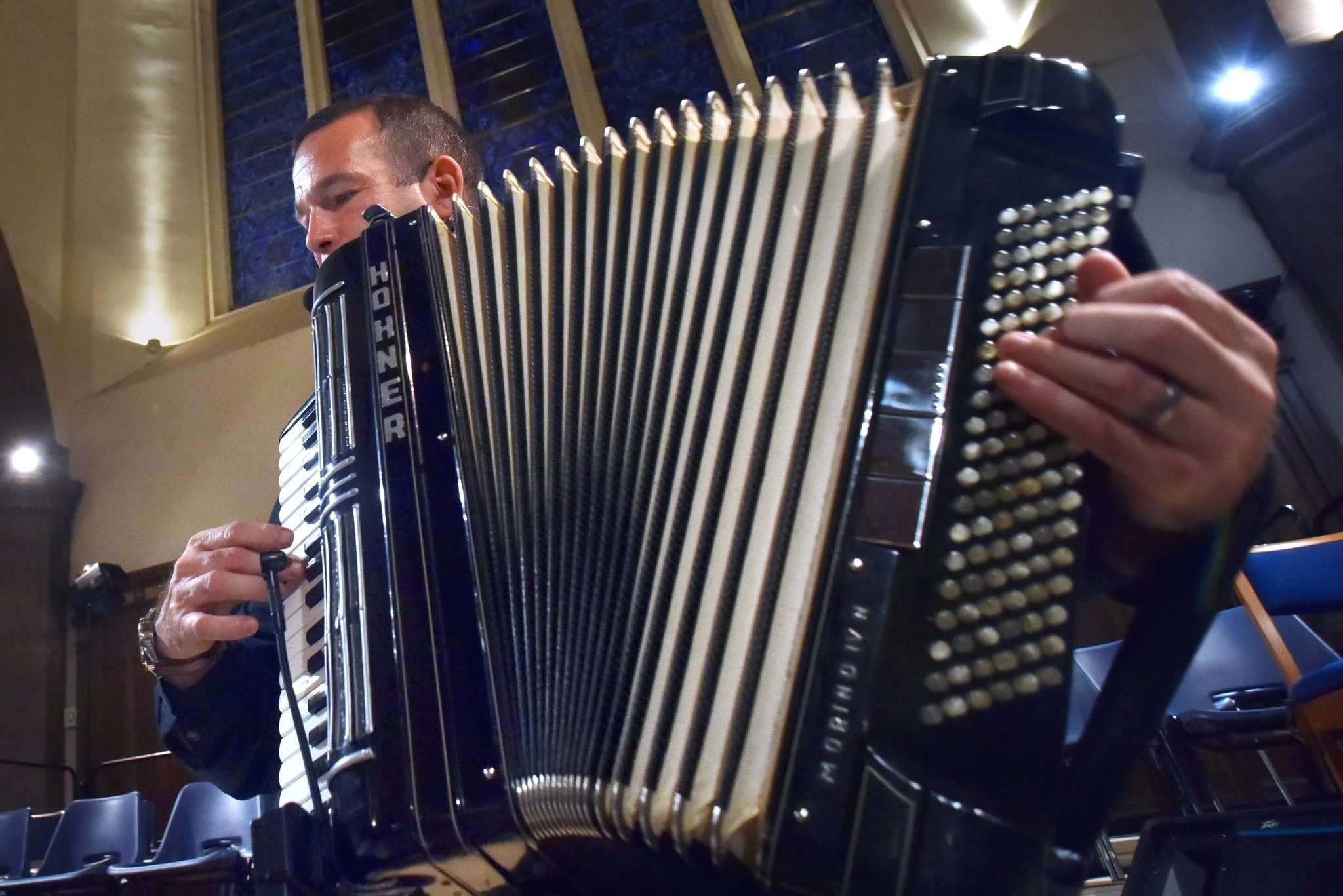 A man playing an accordion indoors, with a background of windows with blinds and purple-blue trees outside.  Clappy Doo Ceilidh band Scottish Ceilidh band Scottish music