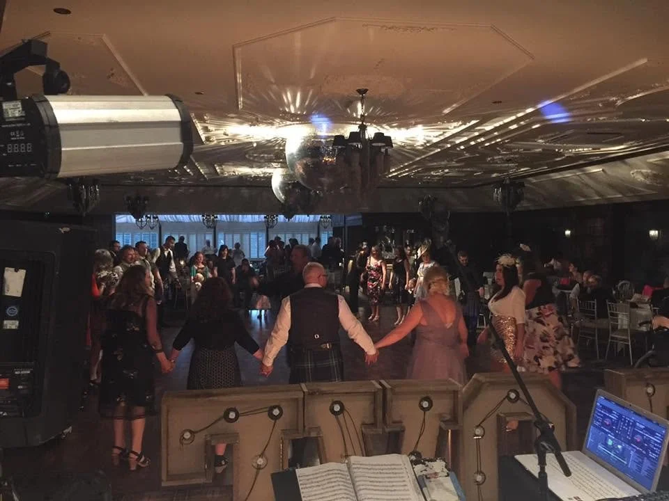 People dancing in a dimly lit ballroom with a DJ booth in the foreground, a disco ball hanging from the ceiling, and a group of friends holding hands in a circle.  Clappy Doo Ceilidh band Scottish Ceilidh band Scottish music
