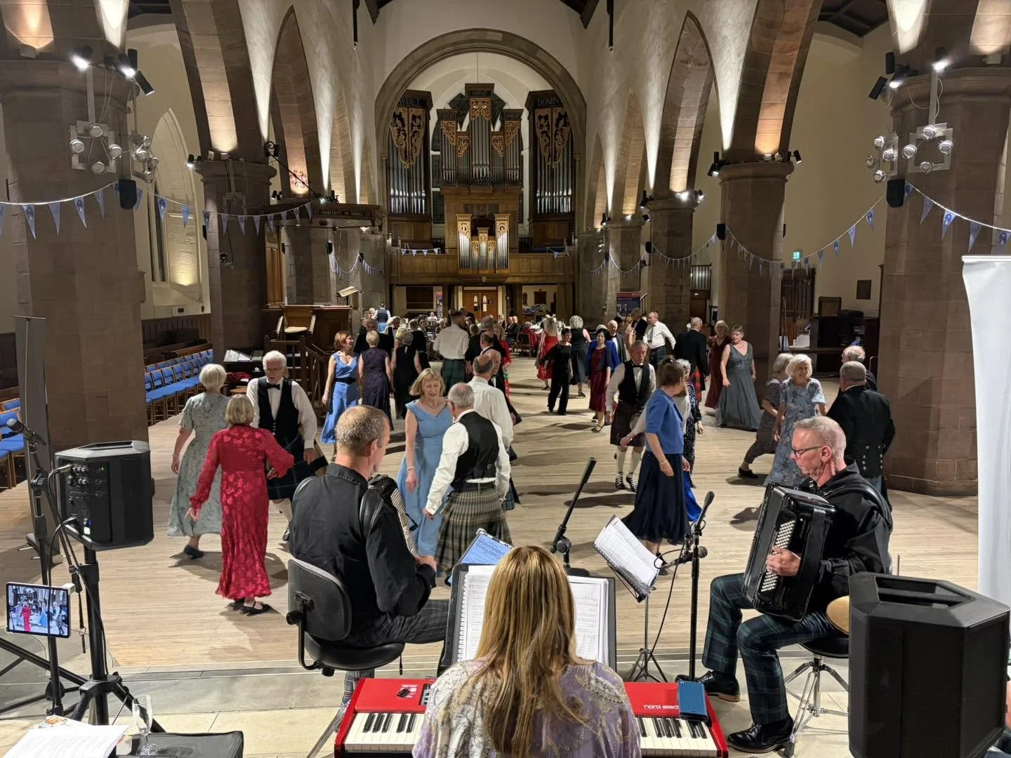 People dancing in a large hall with a live band playing music. The hall has high arched ceilings, stone walls, and an organ at the back. Clappy Doo Ceilidh band Scottish Ceilidh band Scottish music