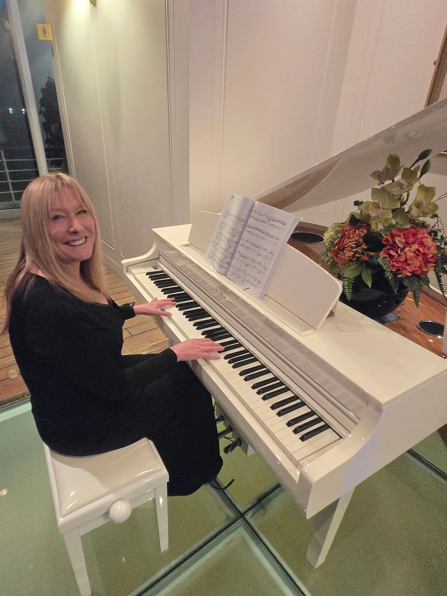 A woman with blonde hair wearing a black dress is playing a white grand piano indoors. There is sheet music on the piano, and a colorful bouquet of flowers is placed on top.  Clappy Doo Ceilidh band Scottish Ceilidh band Scottish music
