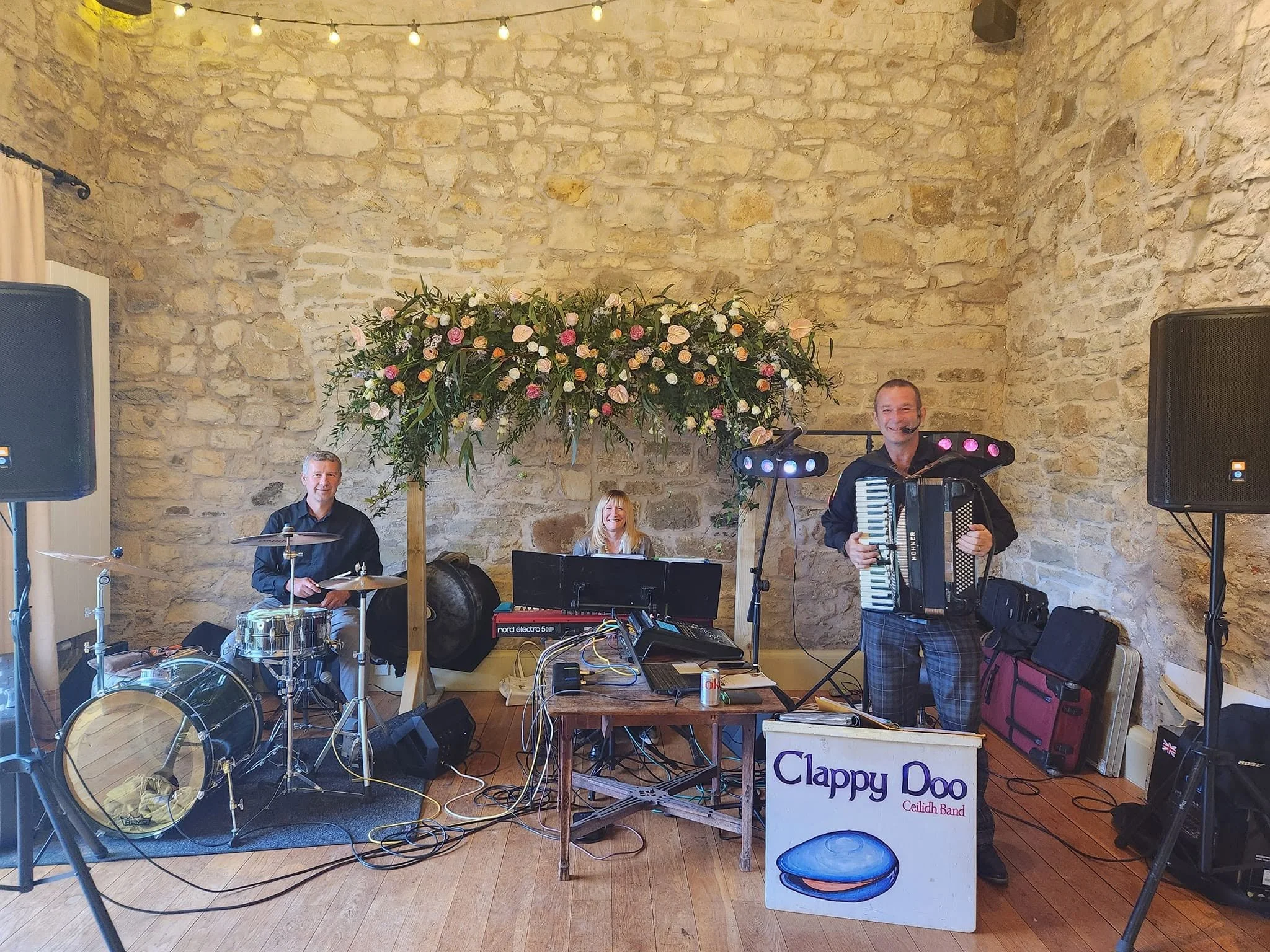 Musicians performing at a live event in a venue with stone walls and string lights, including a drummer, a woman at the keyboard, and a man playing an accordion. There is a flower arrangement and audio equipment visible.