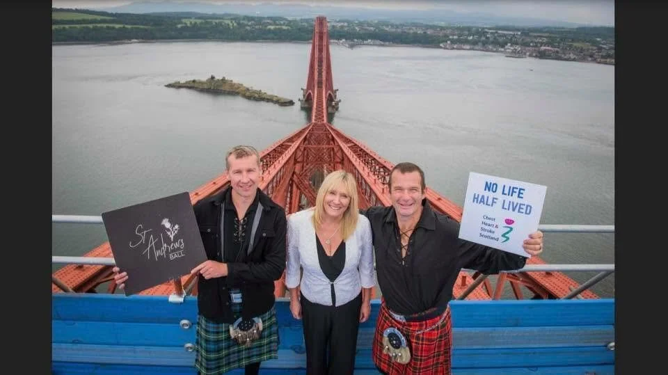 Three people standing on a bridge over a body of water, holding signs. The person on the left is holding a sign that says 'St. Andrews Ball'. The person on the right is holding a sign that says 'No Life Half Lived,' with a heart and Scottish flag sym