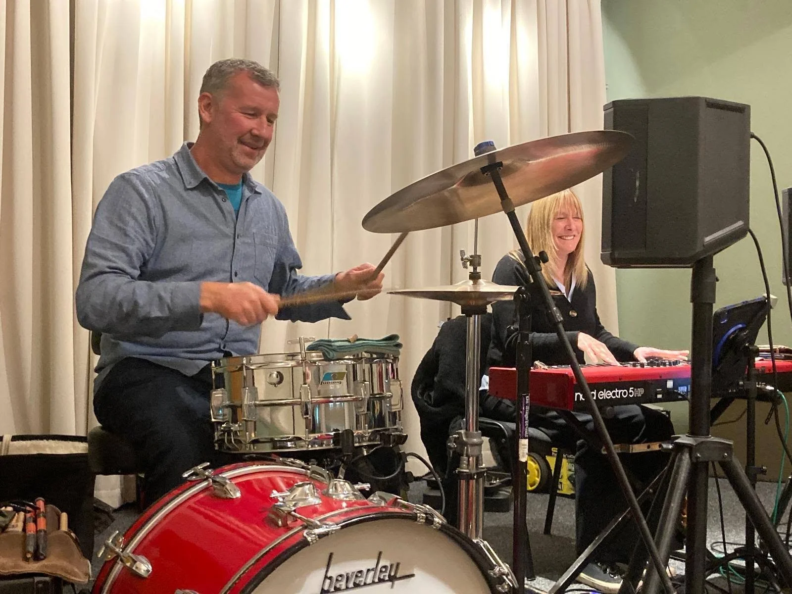 Two musicians, a man and a woman, performing at an indoor event. The man is playing drums, and the woman is playing a red keyboard. They are smiling and the background has a cream-colored curtain.  Clappy Doo Ceilidh band Scottish Ceilidh band 