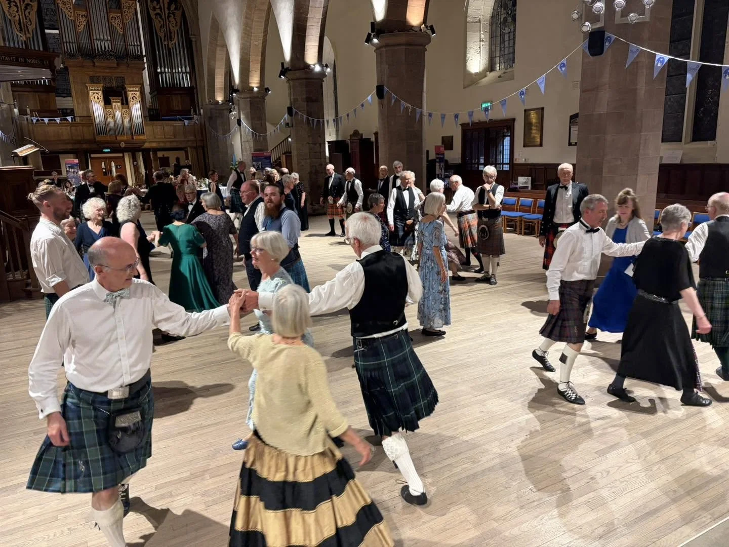 People dancing in pairs inside a large hall with tall arches and an organ in the background, some wearing traditional Scottish clothing like kilts. Clappy Doo Ceilidh band Scottish Ceilidh band Scottish music